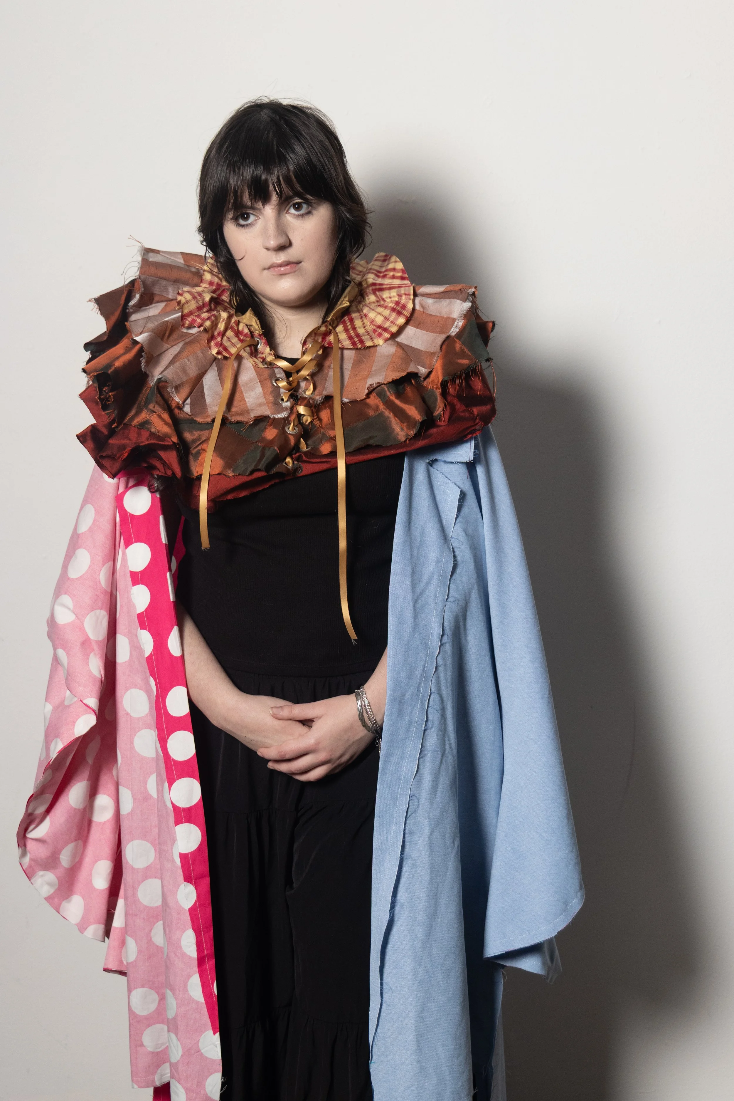 A woman wearing a multi-layered, eclectic outfit with various fabrics, patterns, and textures, standing against a plain wall.