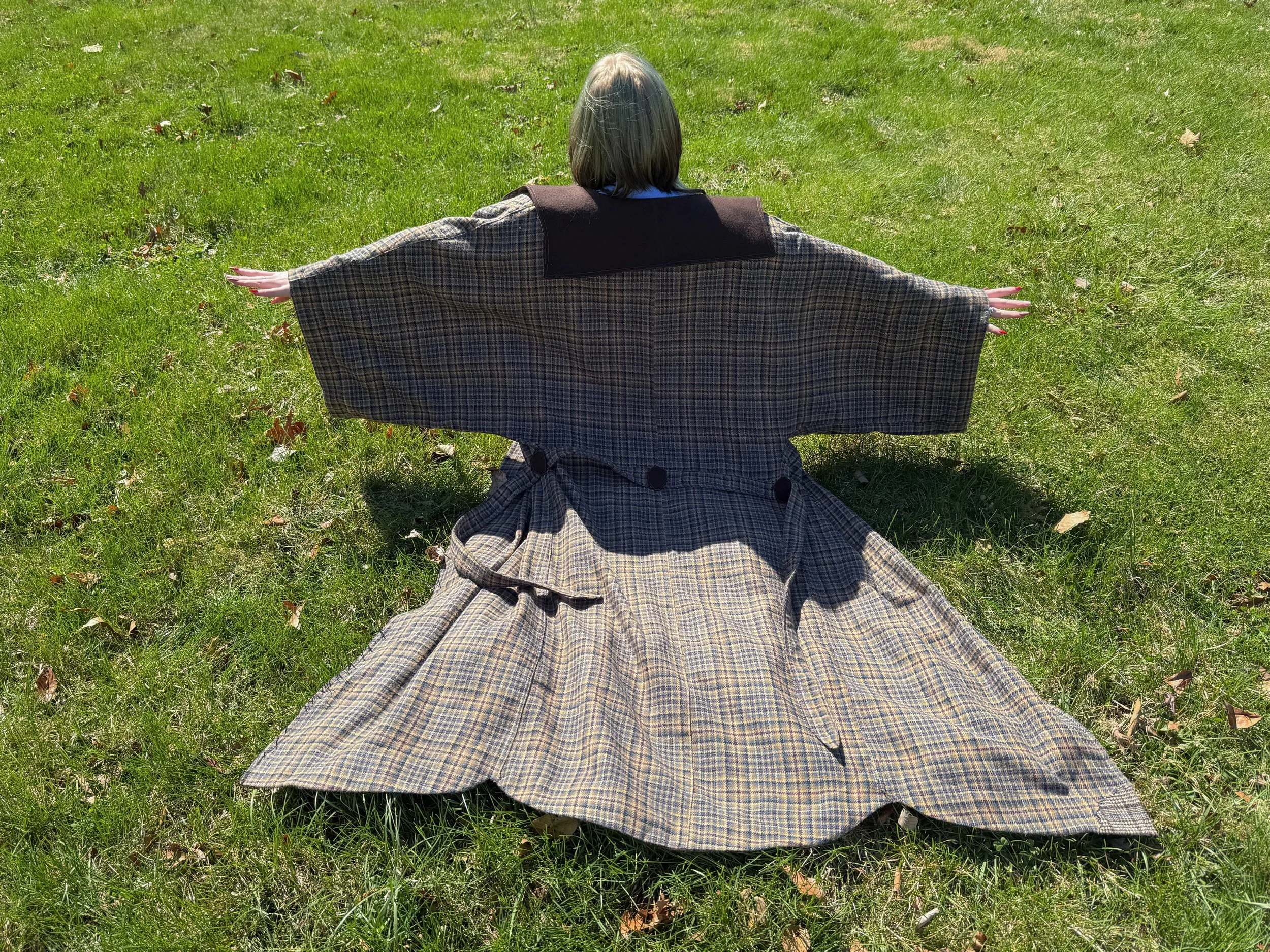 Person with gray hair sitting on grass with arms outstretched, wearing a long beige and brown plaid coat with wide sleeves and a black collar.
