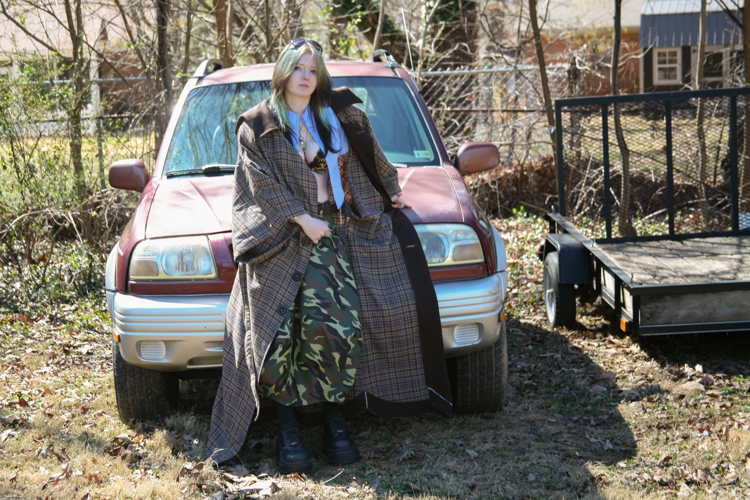 A young woman with blue hair, wearing an oversized plaid coat, camouflage skirt, and black boots, leaning against the hood of a maroon sedan parked on a grassy area with leafless trees and a trailer to her right.