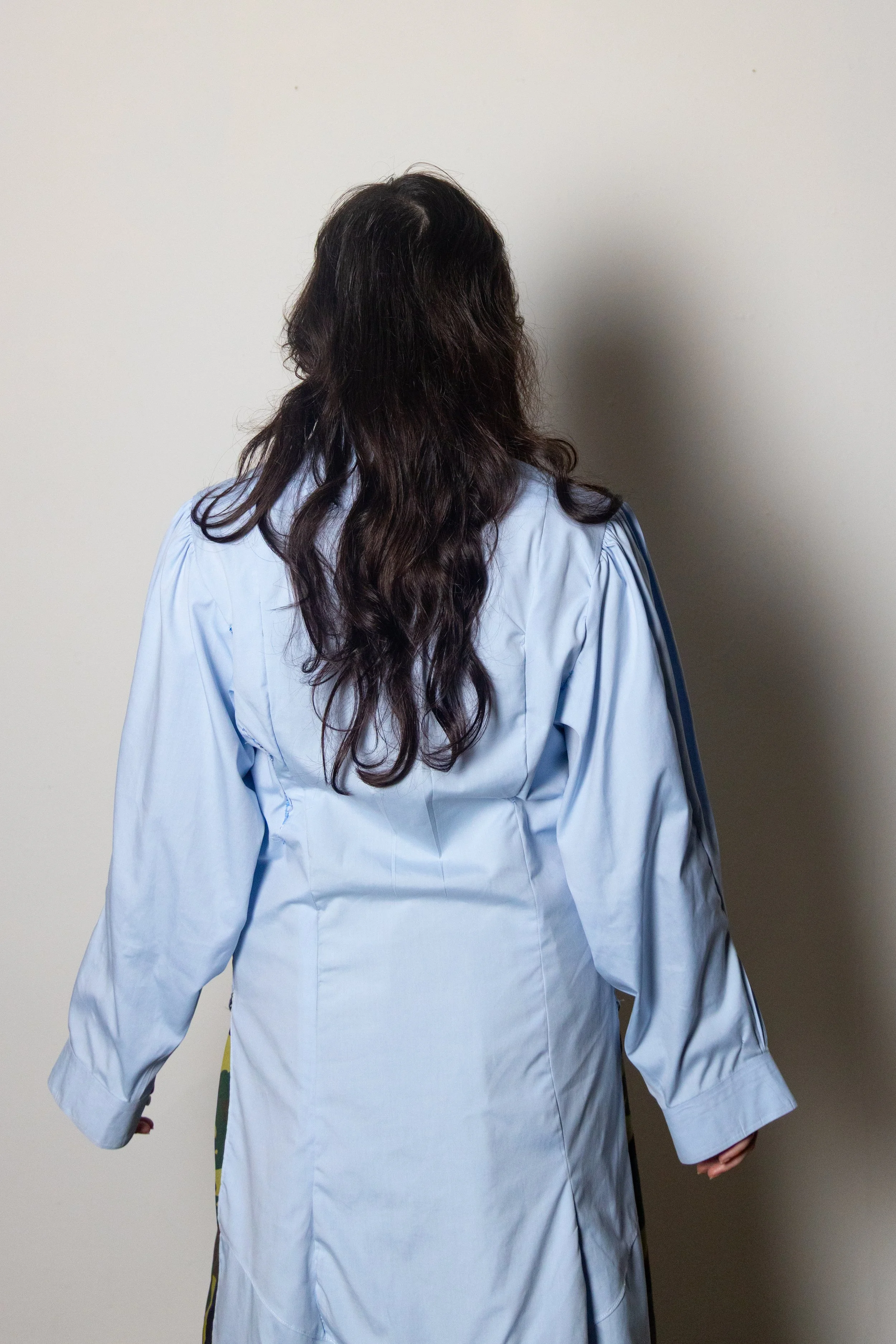 Back view of a woman with long, wavy dark hair wearing a light blue medical gown standing against a plain wall.