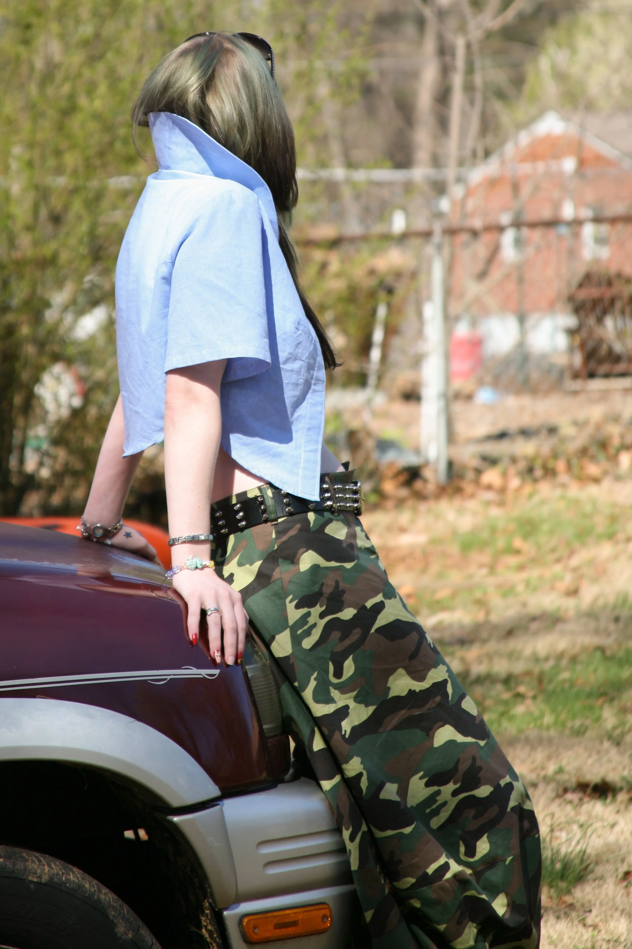 Person with long dark hair wearing a blue cropped shirt and camouflage pants, leaning against a maroon vehicle outdoors on a sunny day.