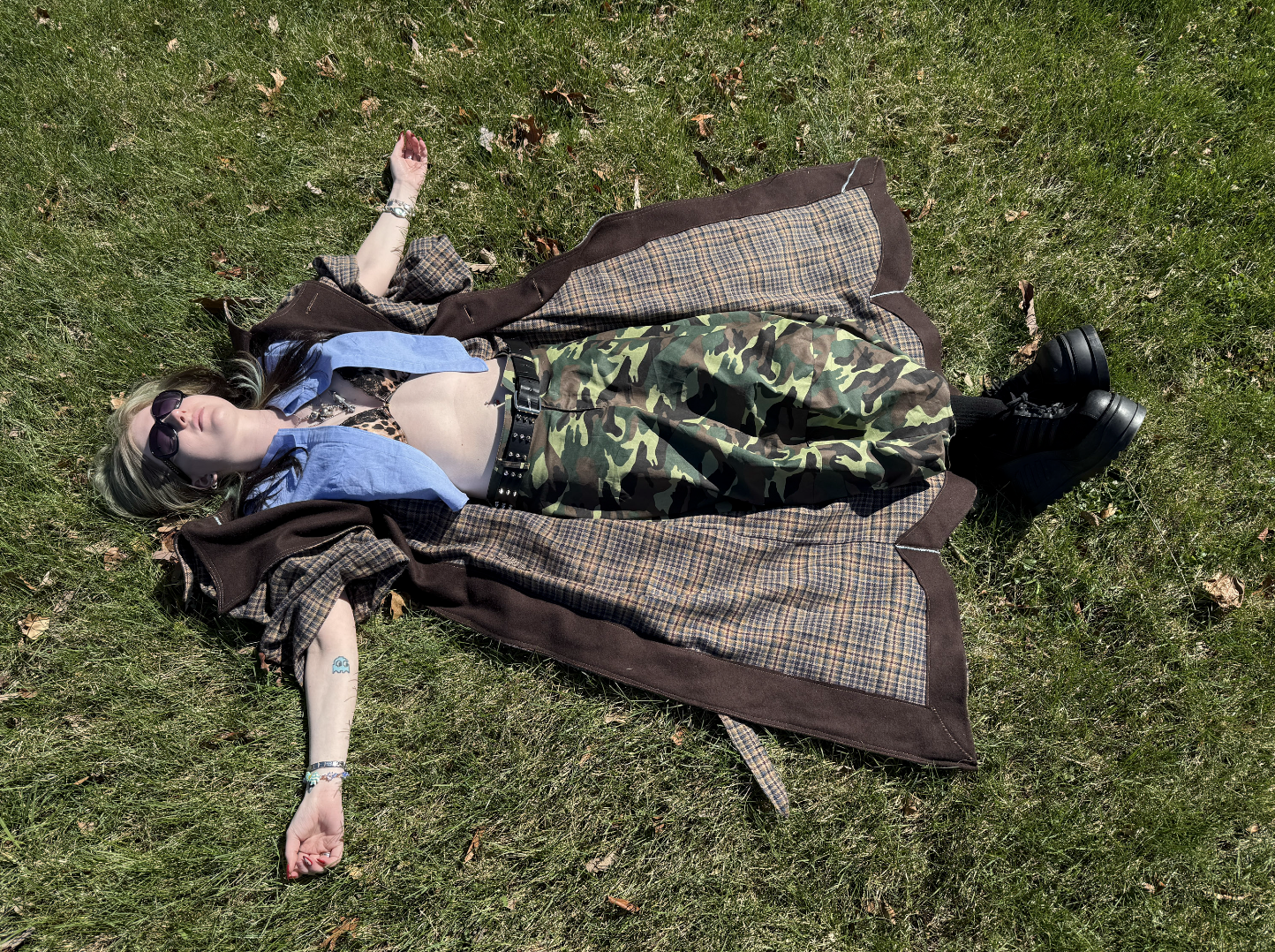 A woman lying on grass outdoors, dressed in a Frankenstein-themed shirt, camouflage pants, wearing sunglasses, with a patterned coat draped over her. She has bracelets on her wrist and is wearing black platform shoes.