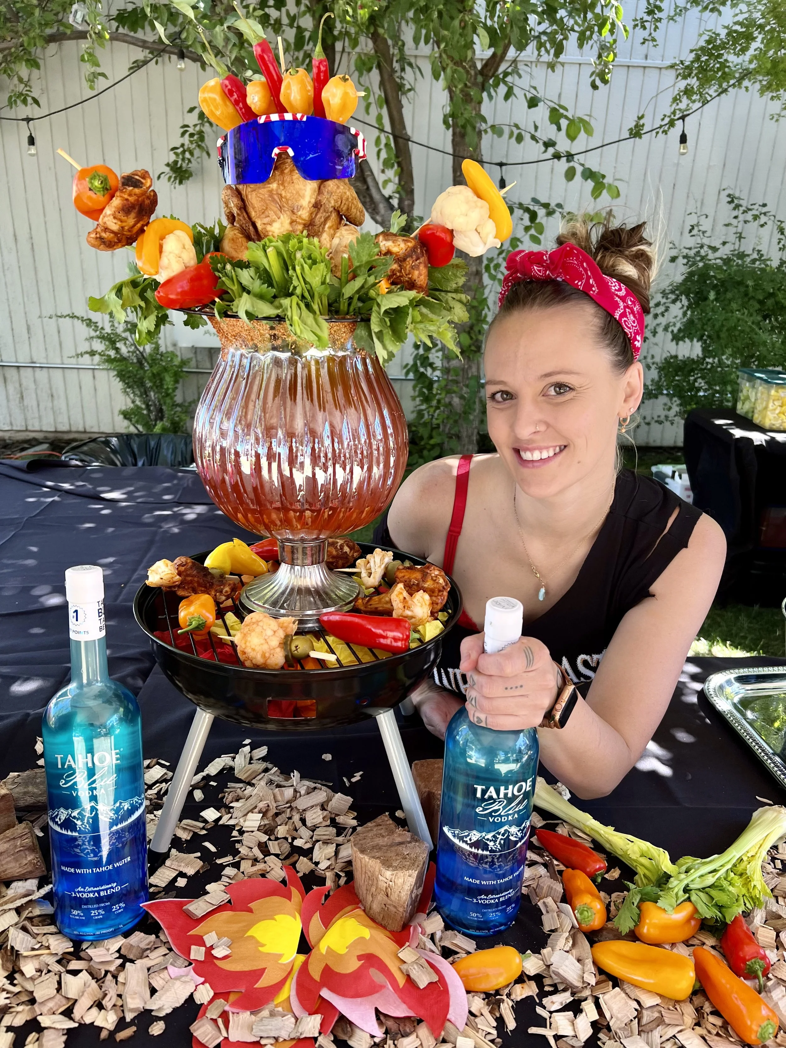 A woman with a red bandana on her head smiling while sitting at an outdoor table with a festive display of food and drinks. There is a small charcoal grill with various vegetables and cooked chicken, and a large glass vase with a roast chicken decorated with colorful peppers and sunglasses on top. Two bottles of Tahoe blue vodka are on the table, surrounded by fire-themed paper decorations, celery stalks, and peppers, all on a black tablecloth with wood chip decorations and greenery in the background.