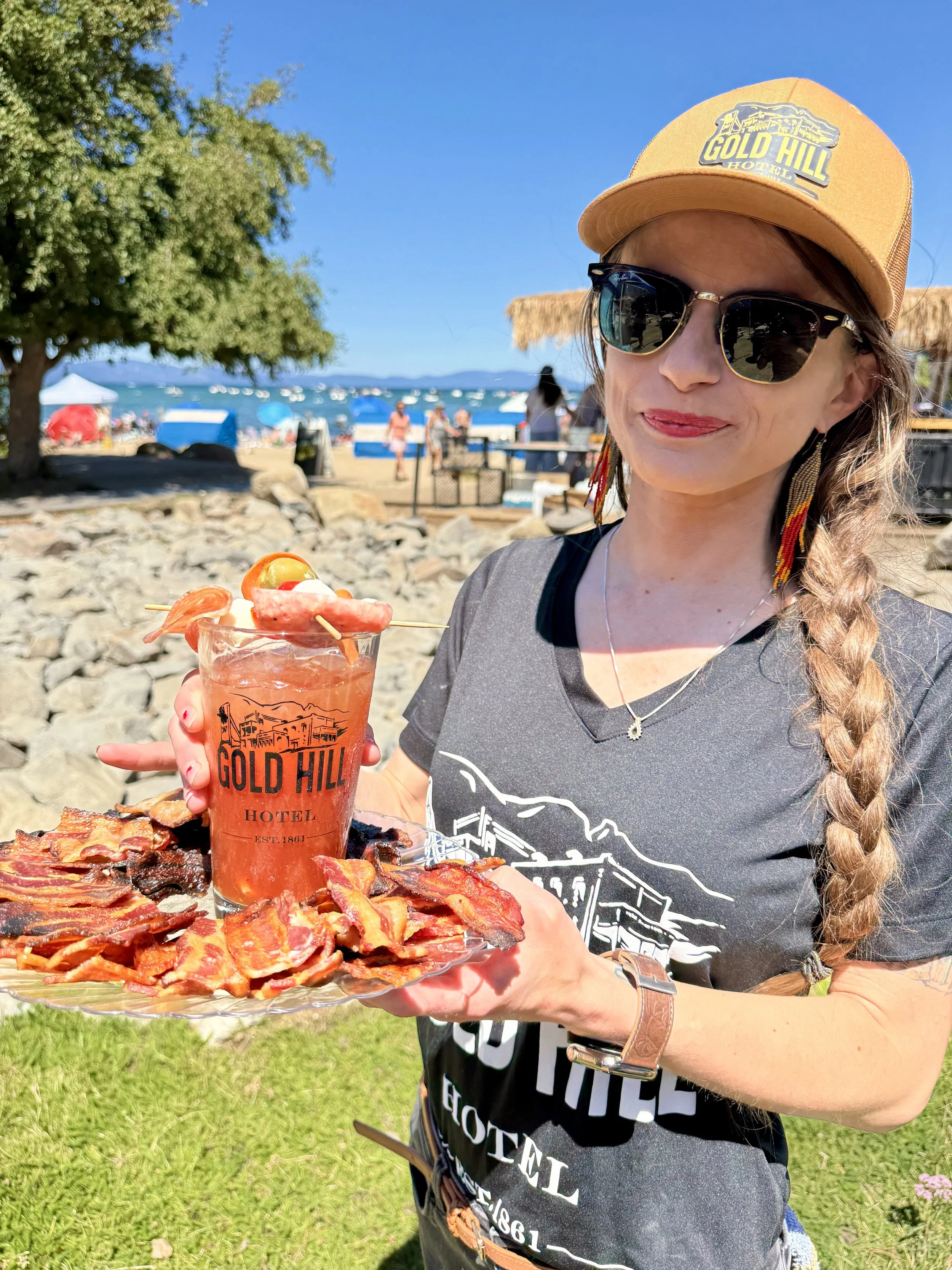 A woman at a beach holding a tray of bacon and a drink in a tall glass with a straw, garnished with fruit. She is wearing sunglasses, a yellow hat with "GOLD HILL HOTEL" embroidered on it, a gray T-shirt, and jewelry. In the background, there are people on the beach, boats in the water, and a large tree.