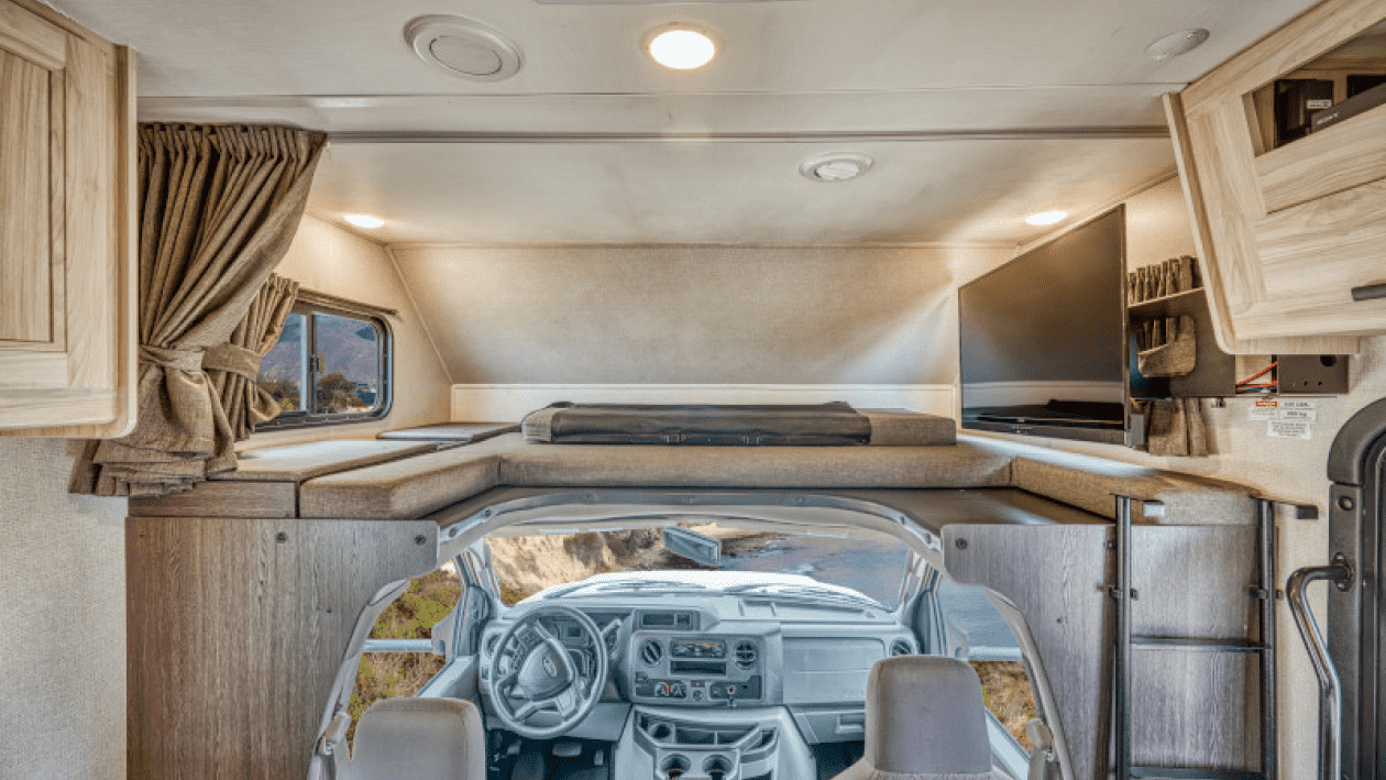 Interior de una vivienda móvil con cama arriba, ventana, televisión y cocina, vista desde la cabina del conductor.