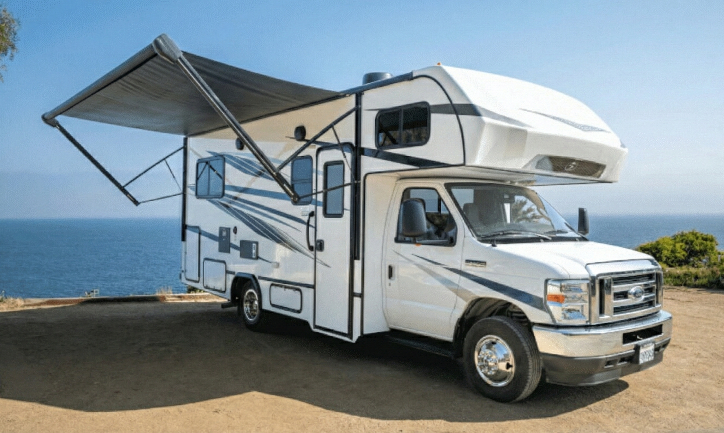 Camioneta tipo motorhome blanca con toldo extendido, estacionada cerca del mar en un área de tierra.