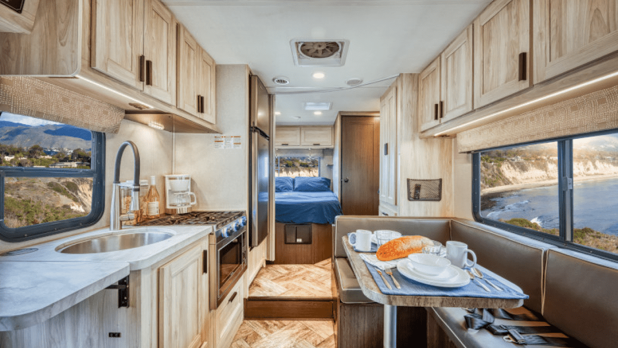Interior de una casa rodante con cocina, comedor y cama con vista a la playa y acantilados.