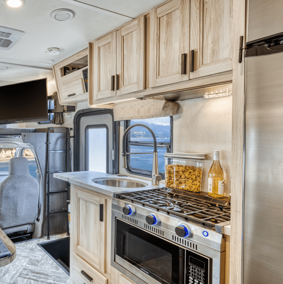 Cocina compacta dentro de una casa rodante, con estufa, fregadero, armarios de madera y una ventana que muestra un paisaje monta