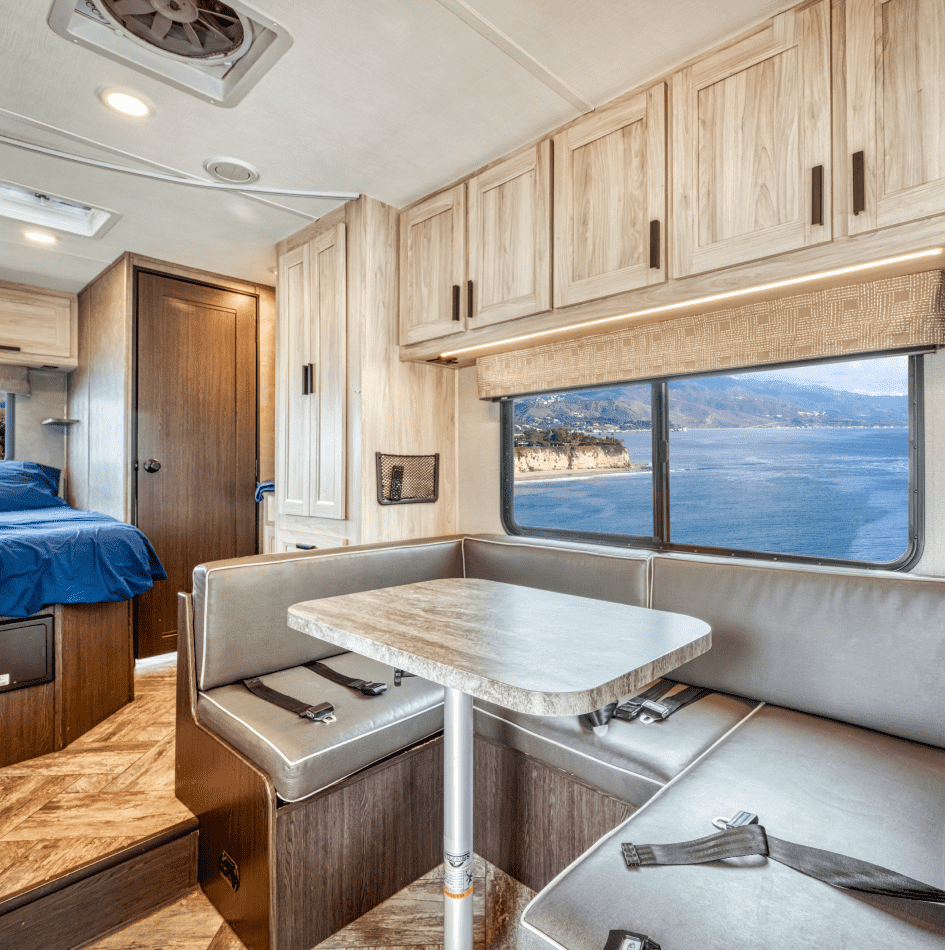 Interior de un bote o casa rodante con vista al mar, con mesa y bancos en la sala.