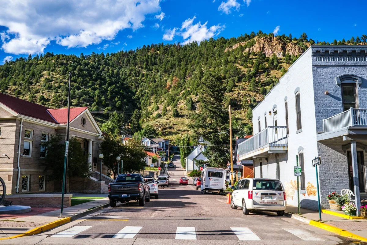 Vista de un callejón en un pueblo con casas, autos estacionados, árboles y una colina o montaña con vegetación en el fondo, bajo un cielo azul con nubes.