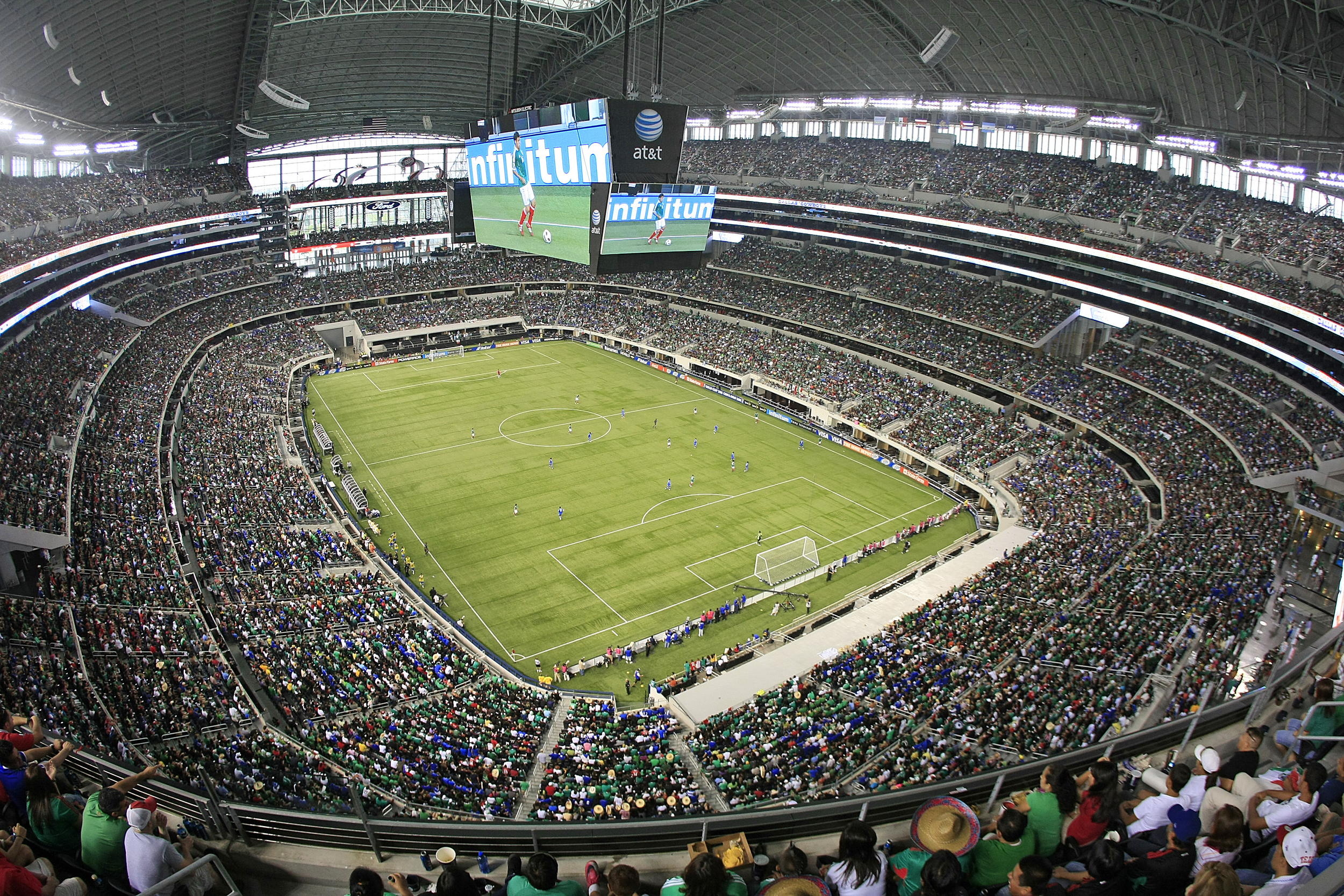 Vista panorámica de un estadio lleno de espectadores durante un partido de fútbol, con un campo verde y una pantalla gigante mostrando jugadores en acción.