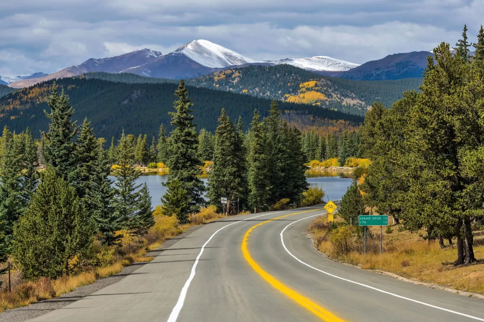 Carretera en un paisaje montañoso con árboles, un lago y picos nevados en el fondo.