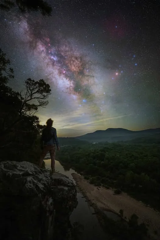 Persona de espaldas observando el cielo estrellado y la Vía Láctea desde una salida en la naturaleza, con árboles, montañas y un río en el fondo.