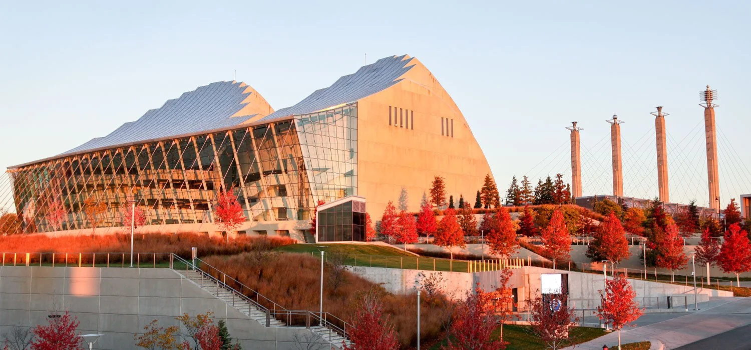 Edificio moderno con ventanas de vidrio y estructura de acero, rodeado de árboles con hojas rojas y un paisaje urbano.