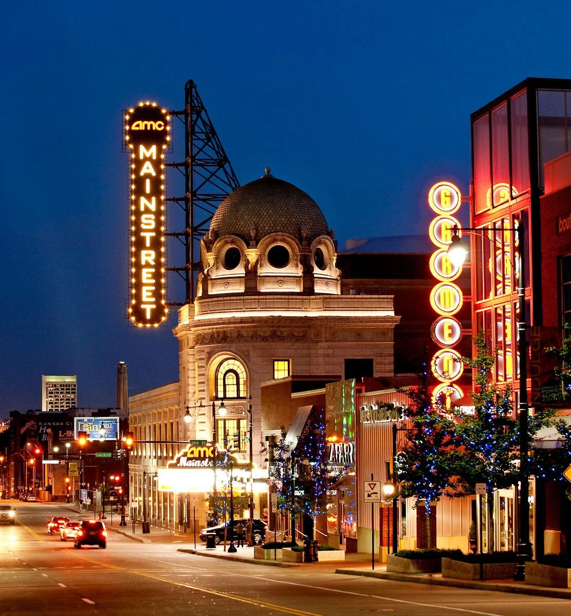 Calles de noche con edificios iluminados, árboles decorados con luces, cartel de cine AMC Mainstreet, y un edificio con cúpula en el centro.
