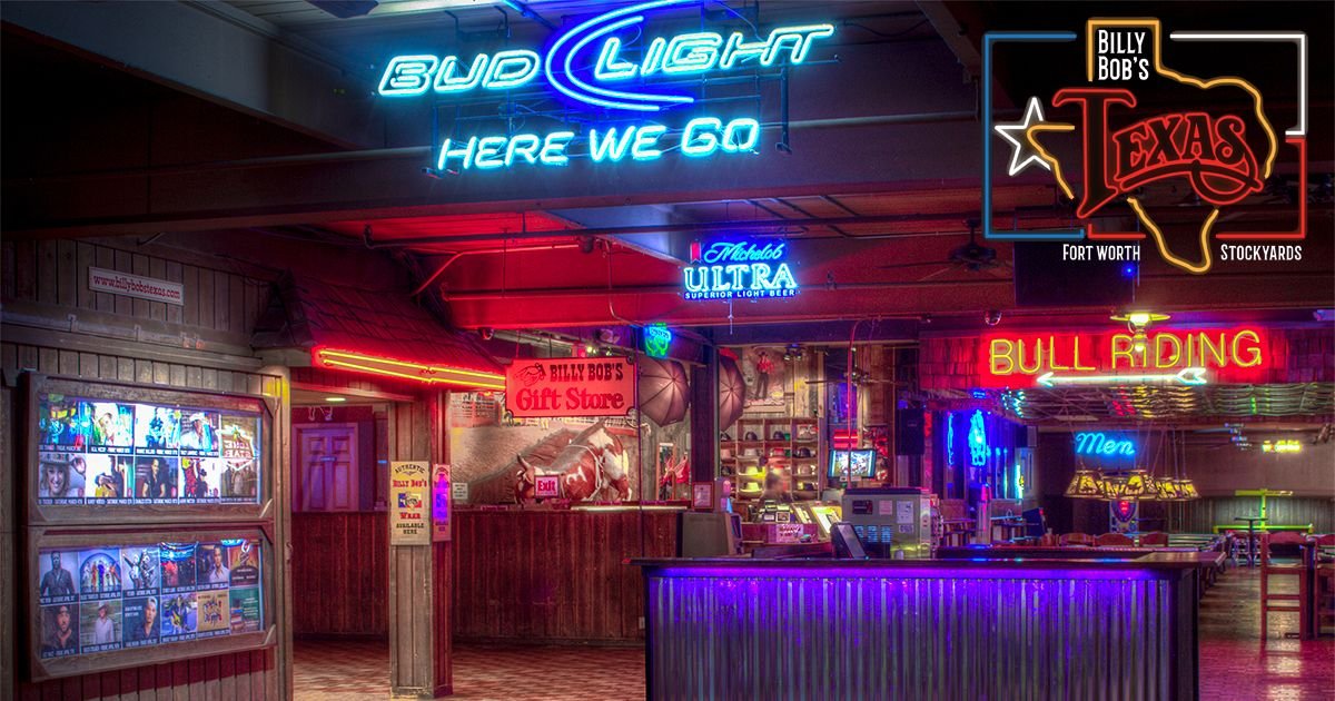 Interior de un bar o salón con carteles de neón, incluyendo uno que dice 'Bud Light Here We Go', otro que dice 'Billy Bobs Gift Store', 'BULL RIDING', y 'Men'. Se puede ver un mostrador con caja registradora y varias señales y decoraciones de neón en las paredes.