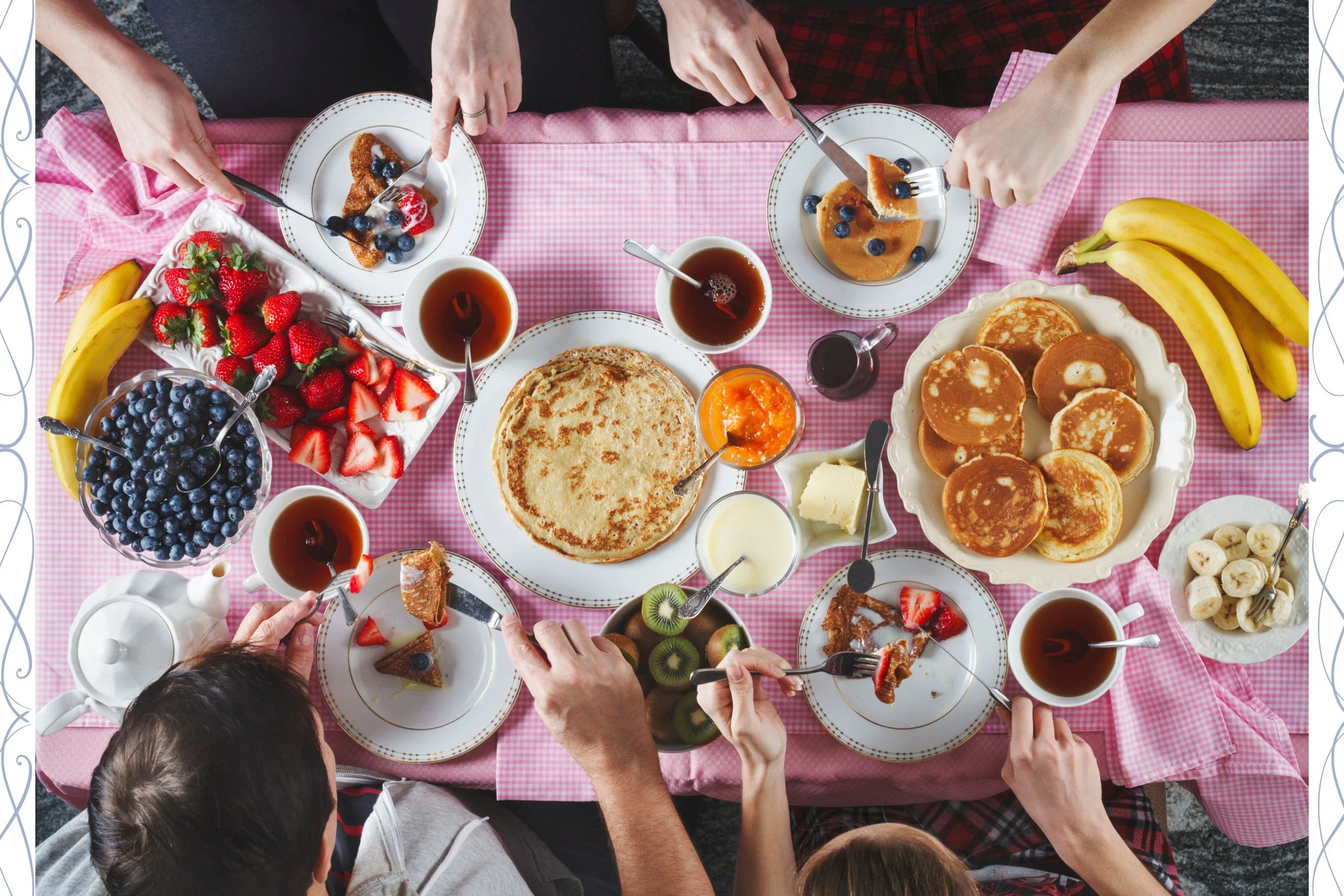 Mesa de desayuno con panqueques, frutas, miel, mantequilla, y bebidas, rodeada de varias personas sirviéndose y comiendo.