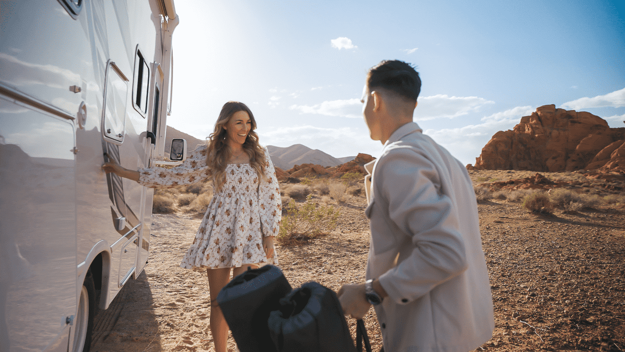 Una mujer y un hombre en un desierto con un autobús de fondo. La mujer está sonriendo y entrando al autobús, mientras que el hombre sostiene una mochila y la observa. El paisaje tiene rocas y montañas, con un cielo despejado.