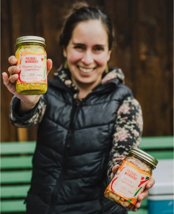 A woman smiling and holding up two jars of homemade preserves, with a wooden fence and a green park bench in the background.