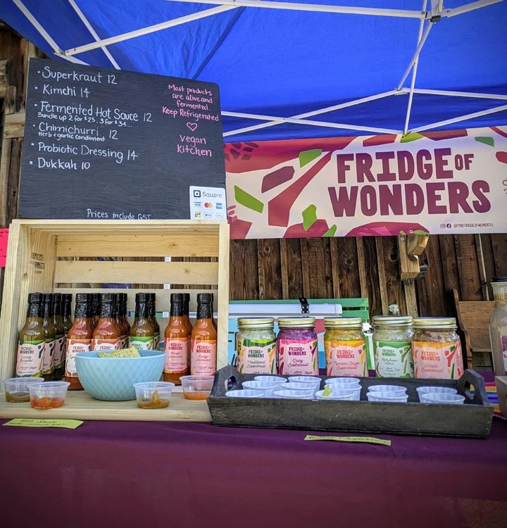 Display of vegan sauces and dressings at a booth named 'Fridge of Wonders' with a menu board listing items like superkraut, kimchi, fermented hot sauce, chimichurri, probiotic dressing, and dukkah, along with jars and bottles of sauces on the table.
