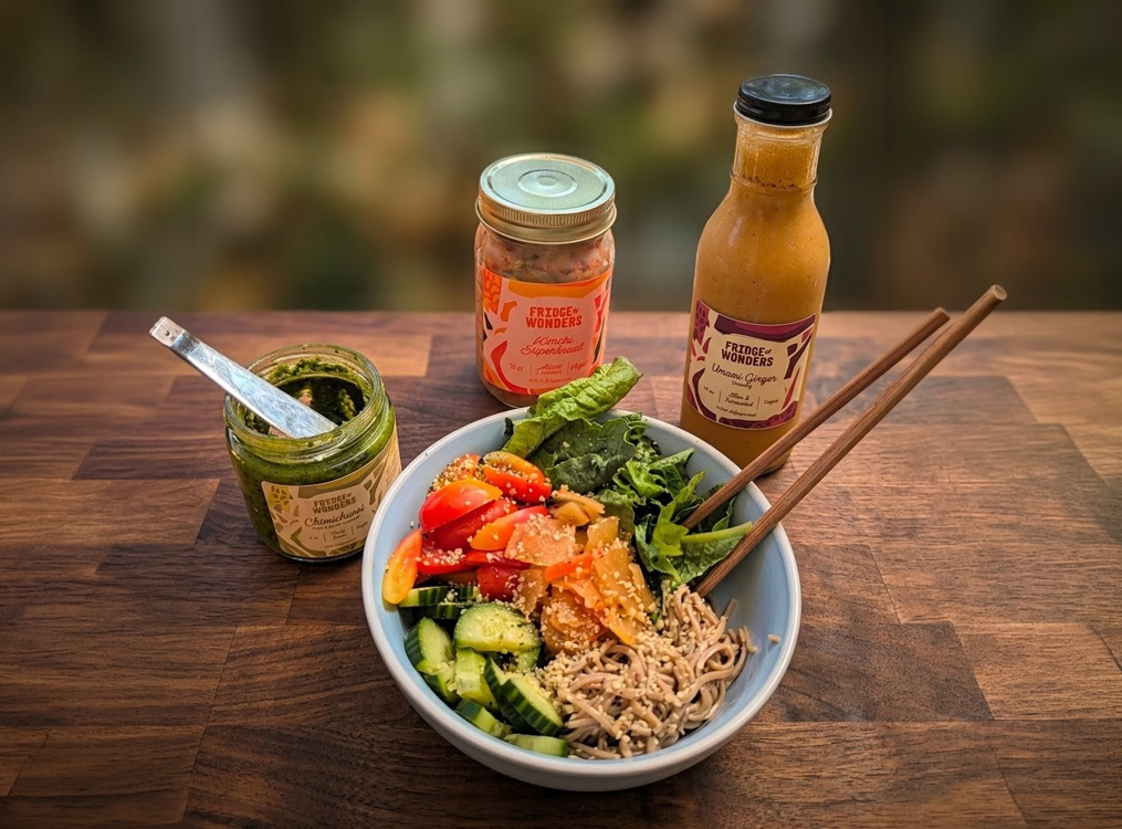A bowl of mixed salad with cucumber, cherry tomatoes, lettuce, shredded carrots, and noodles topped with sesame seeds. Two chopsticks rest on the bowl's edge. Two bottles and a jar of condiments are on the wooden table: a green pesto, a pinkish dressing, and an orange ginger dressing.