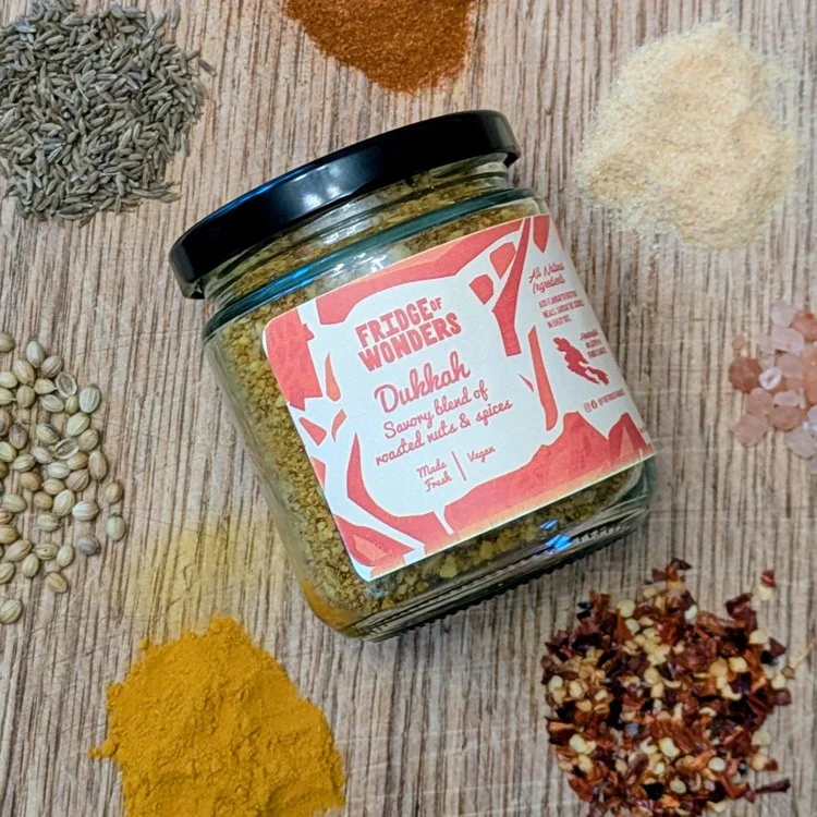 A glass jar labeled "Fridge of Wonders Dukah" on a wooden surface, surrounded by various spices and herbs including turmeric, chili flakes, mustard seeds, ground spices, and pink Himalayan salt.