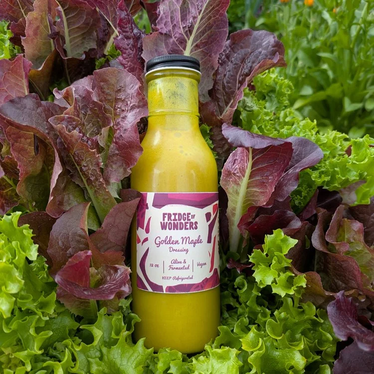 A bottle of Golden Maple Salad Dressing from Fudge of Wonders, placed among red and green leaf lettuce in an outdoor garden.