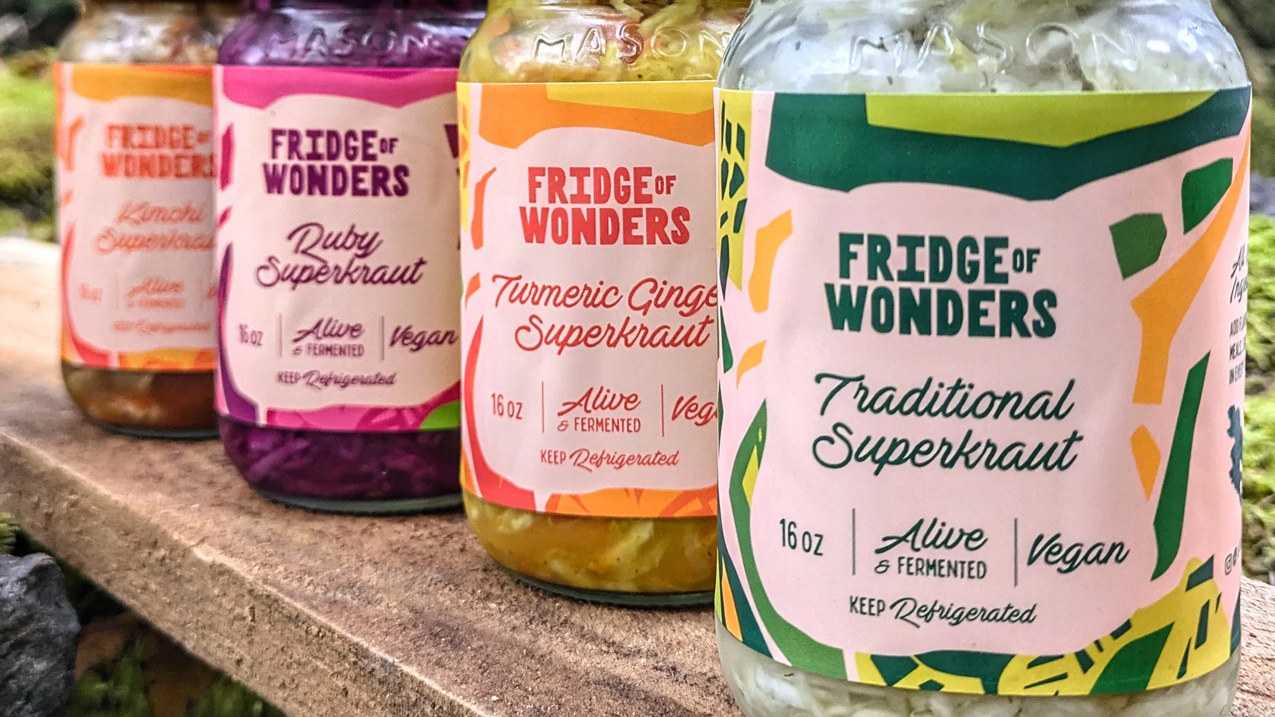 Four jars of superkraut in colorful packaging with different flavors, placed on a wooden surface outdoors. The flavors include traditional, turmeric ginger, ruby, and a partial label for a fourth flavor.