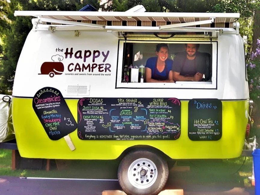 A bright yellow and white food truck named "The Happy Camper" offering homemade sweets and savory snacks, with a menu on black chalkboards and a window with two smiling people inside.