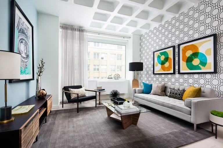 Modern living room with patterned accent wall, white sofa, black armchair, coffee table, colorful abstract artwork, and large window with sheer curtains.