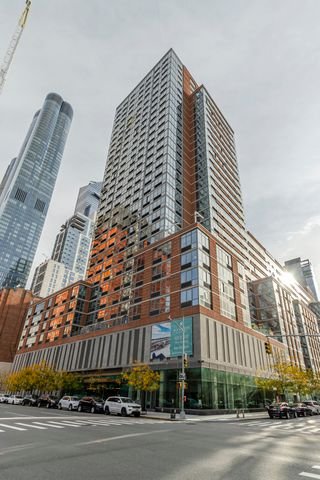 A tall modern skyscraper with a glass facade, featuring a mix of orange, brown, and gray exterior panels, located in an urban cityscape with other high-rise buildings and a street with parked cars and trees.