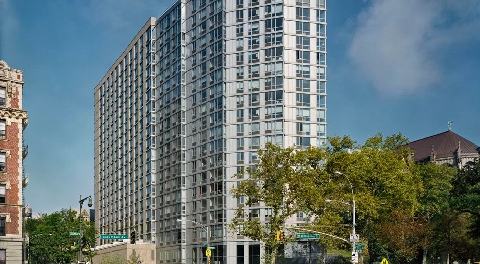 A tall modern high-rise building with a glass exterior in an urban area. There are trees and street signs in the foreground, with a partly cloudy blue sky overhead.