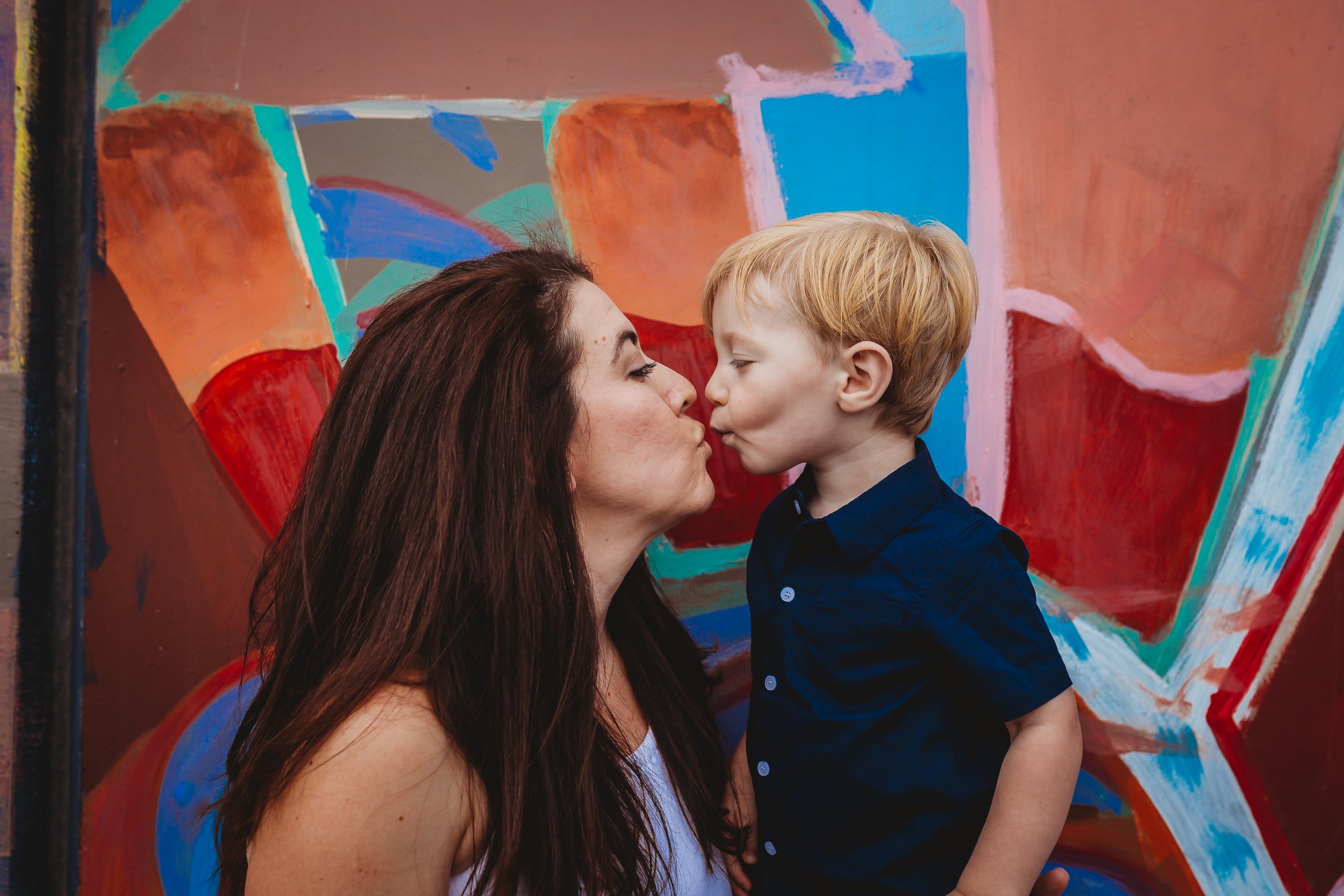 Mommy and Me session located in downtown Colorado Springs, Colorado with a colorful graffiti wall beind them. Toddler and mom are giving each other a silly kiss. 