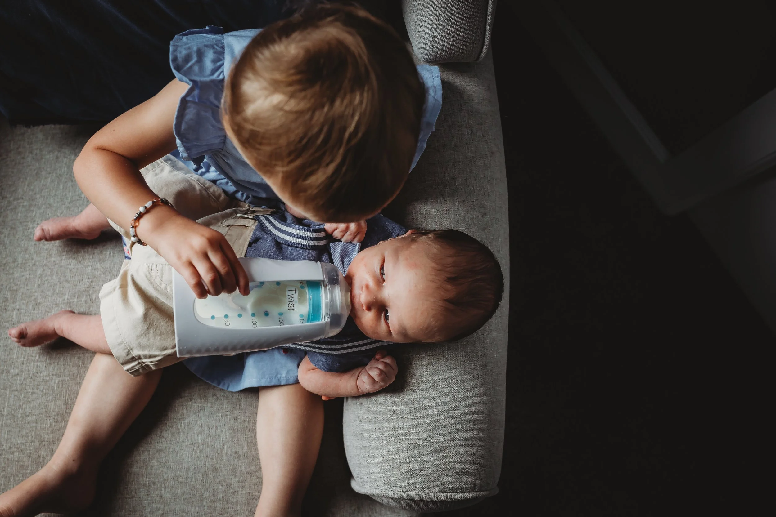 Toddler sister holding newborn brother in a chair in nursery. Natural Light wrapping around them while sister is feeding brother his bottler. Located in Colorado Springs, Colorado. 