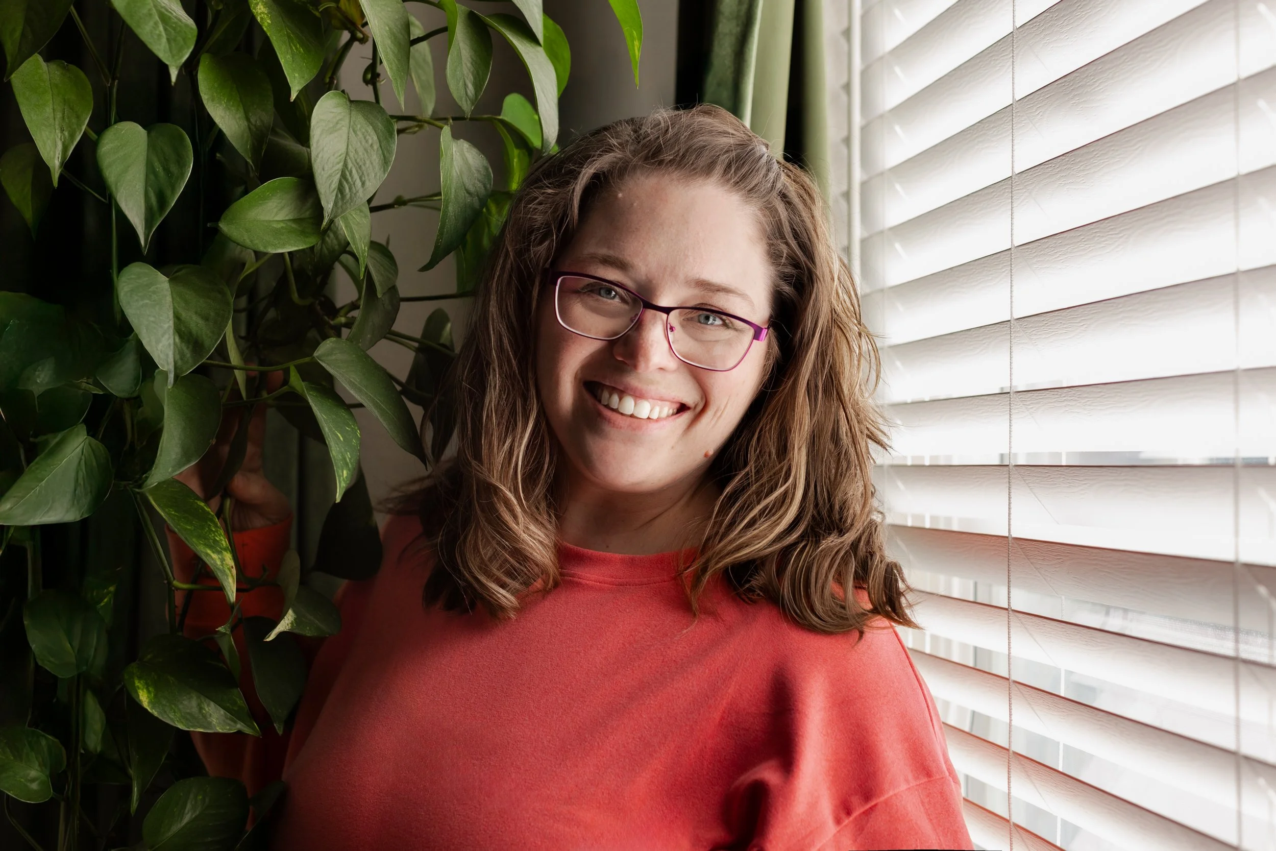 Nikole Aragon from Nikole Aragon Photography in living room with large green plant and window behind her. Self portrait for website.