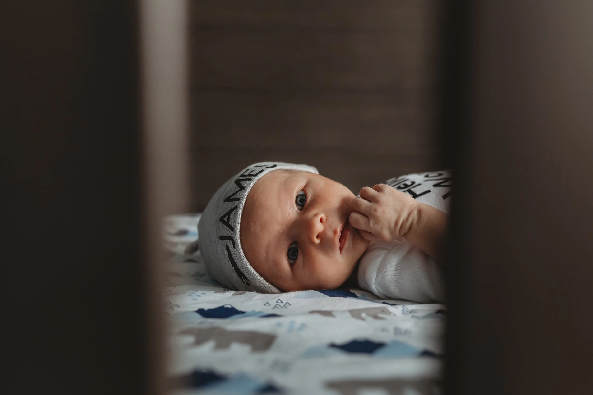 Sweet Newborn boy looking directly into camera through the crib bars in his nursery located in Colorado Springs, Colorado. 
