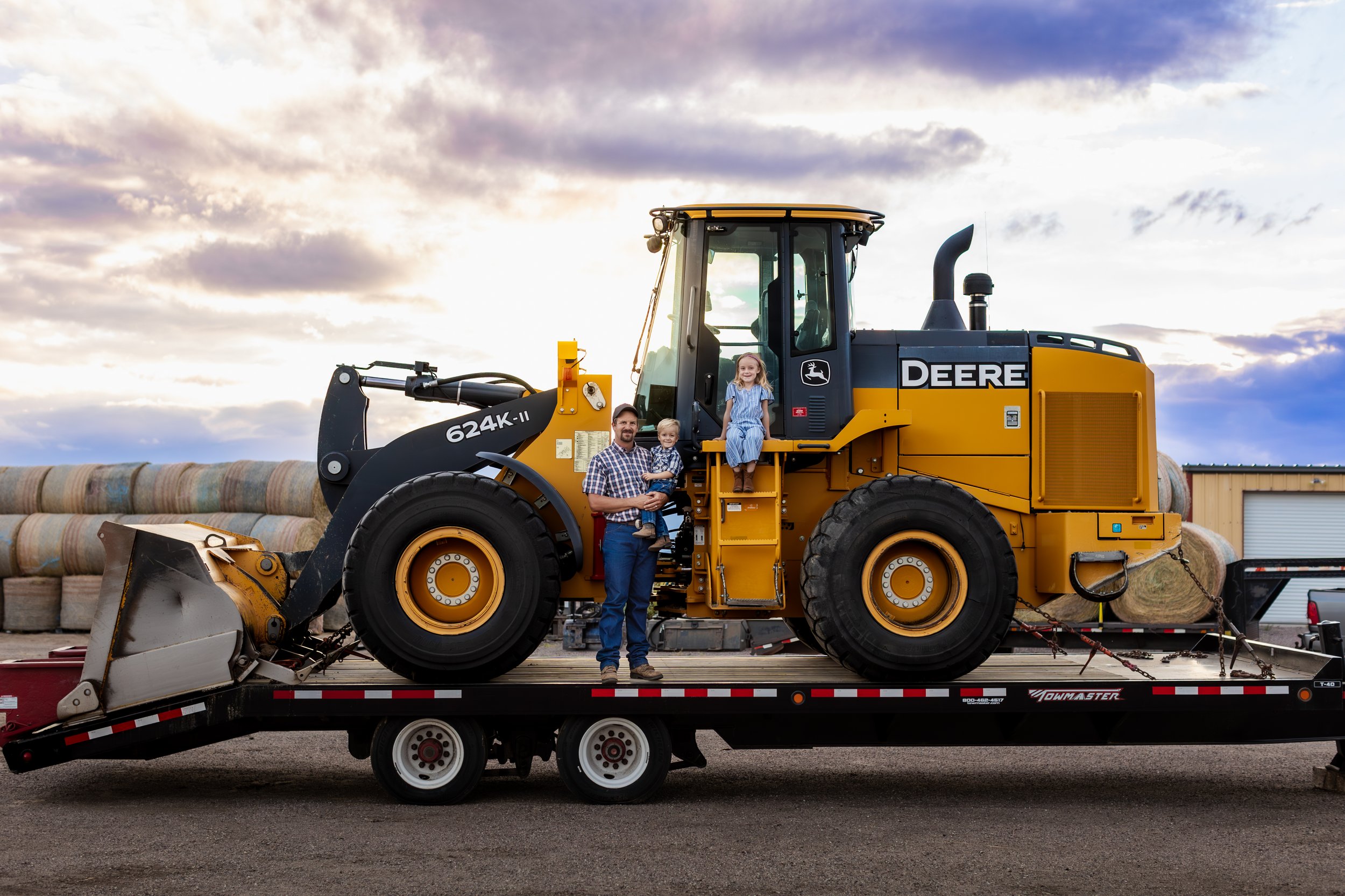 Incredible sunset located in Colorado Springs, Colorado. The farm with hay bails and a large tractor with dad and two children. Toddler boy held by dad and toddler daughter sitting on tractor. 