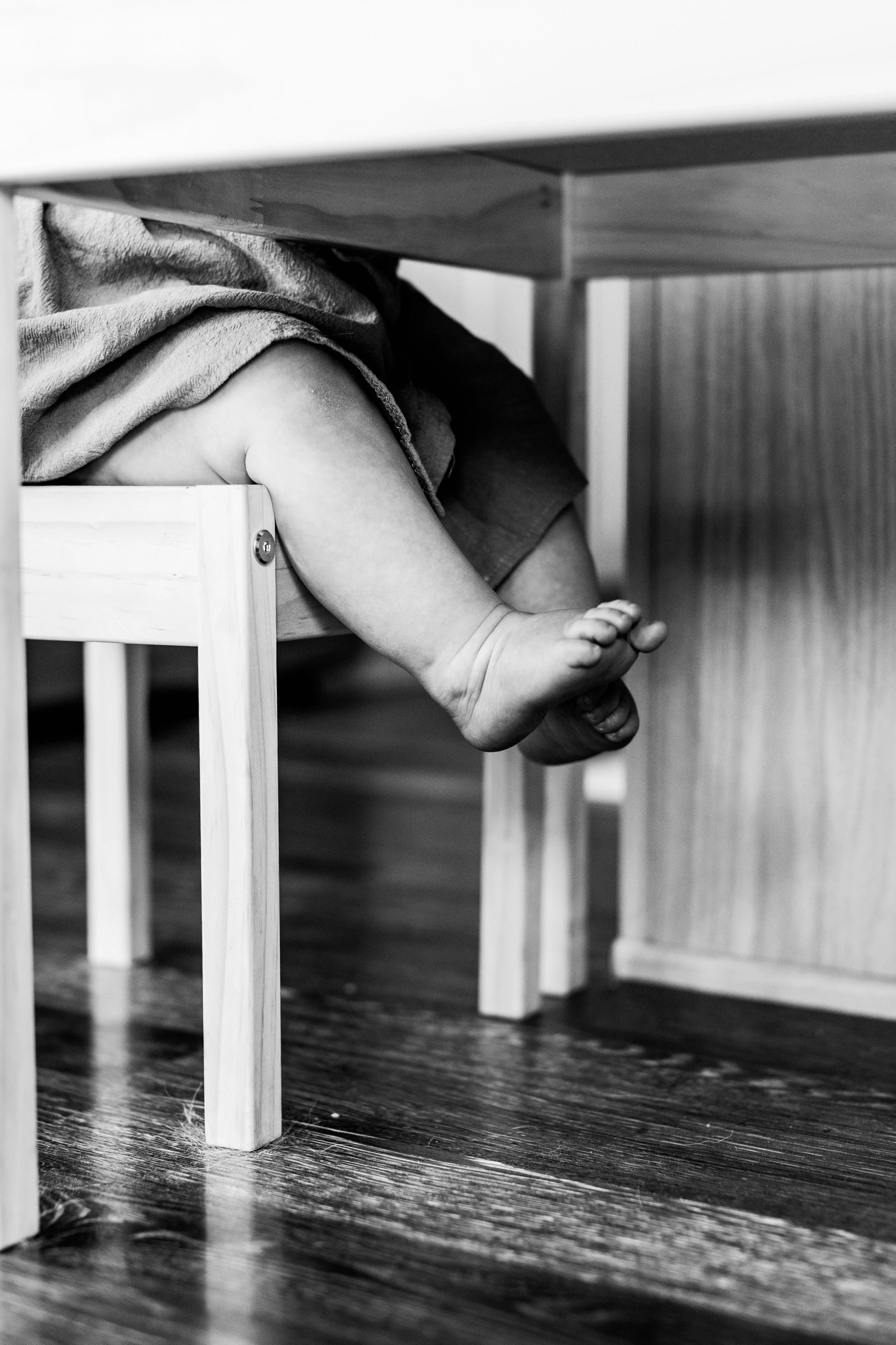 Black and White image of a todler sitting on a chair only showing her feet and legs. Located in home for her first birthday in Colorado Springs, Colorado. 