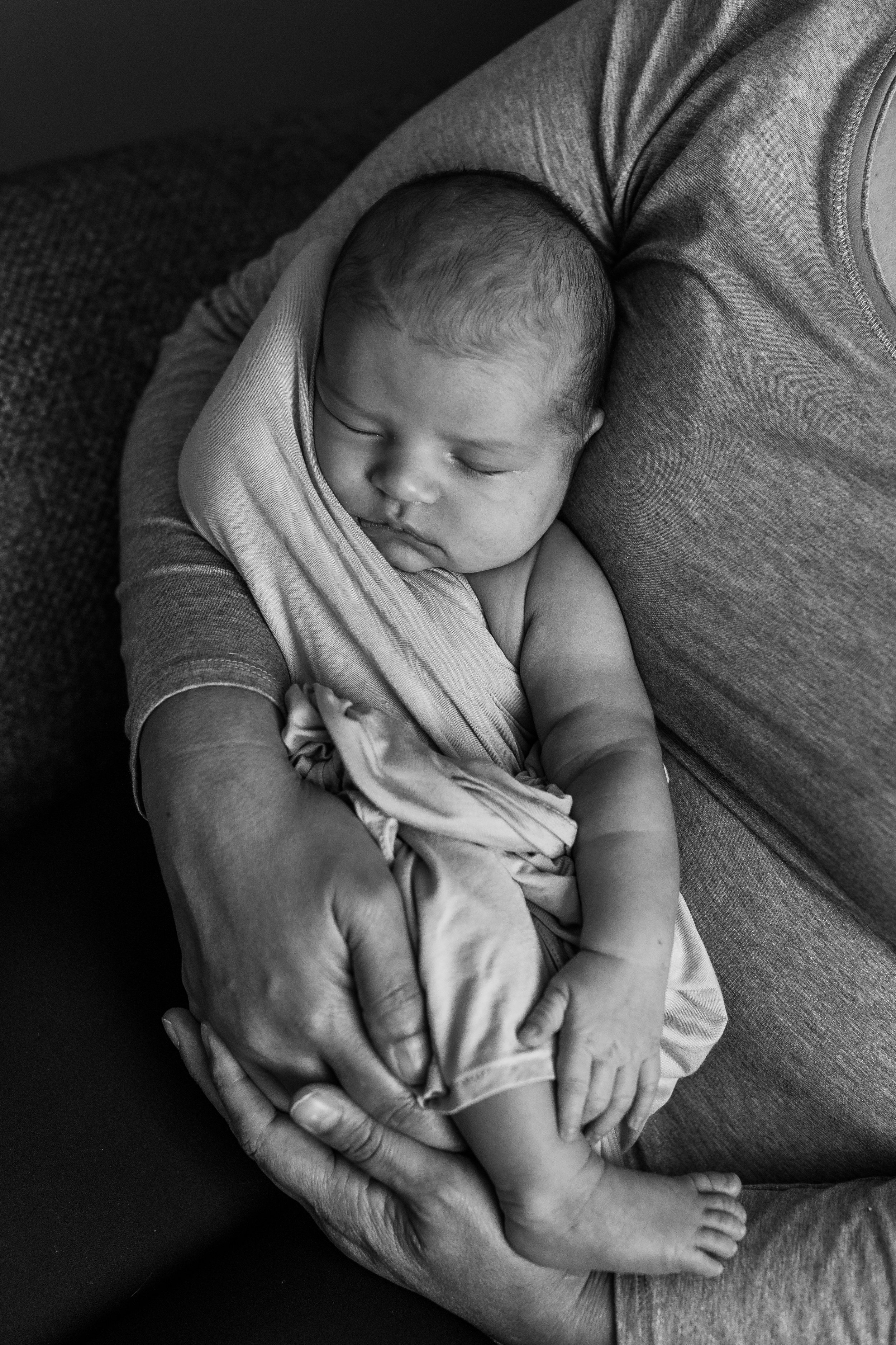 A peaceful newborn baby sleeps cradled in an adult's arms, wrapped in a soft blanket. The black and white image conveys warmth, safety, and tenderness. Comforted in their in home nursery located in Colorado Springs, Colorado.