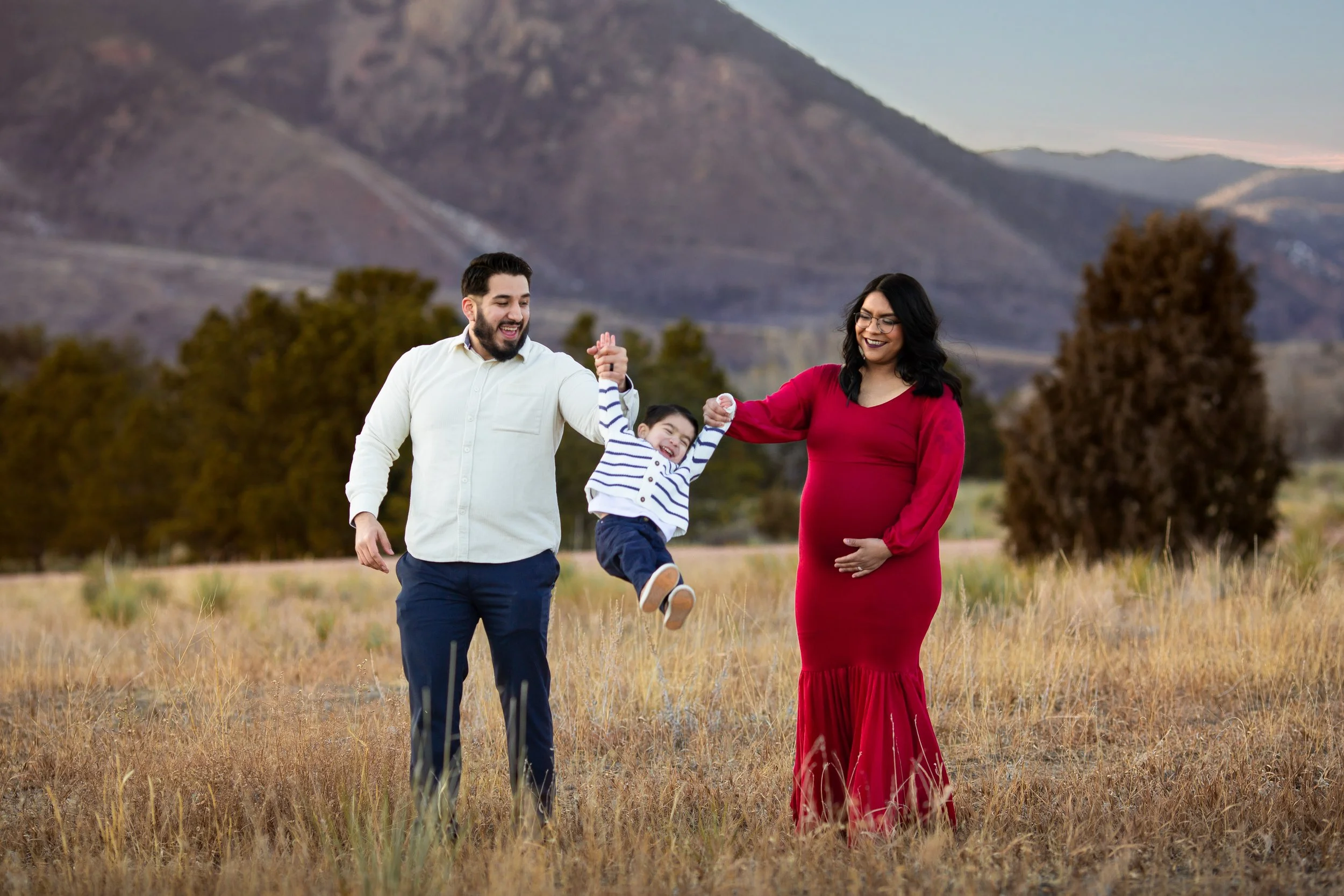 Famiy of three with pregnant mom swinging toddler son between them. The field of tall grasses and mountains with a beautiufl sunset located in Colorado Springs, Colorado. 