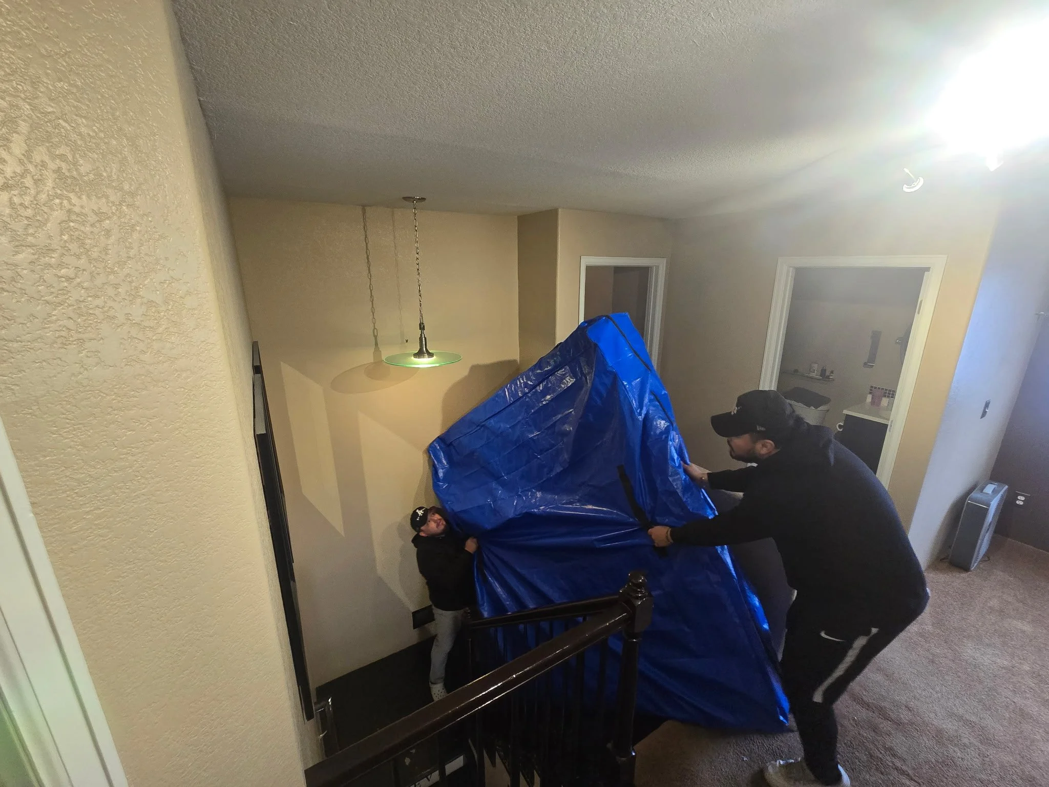 Two individuals are moving a large blue item, possibly furniture or equipment, down a staircase inside a house.