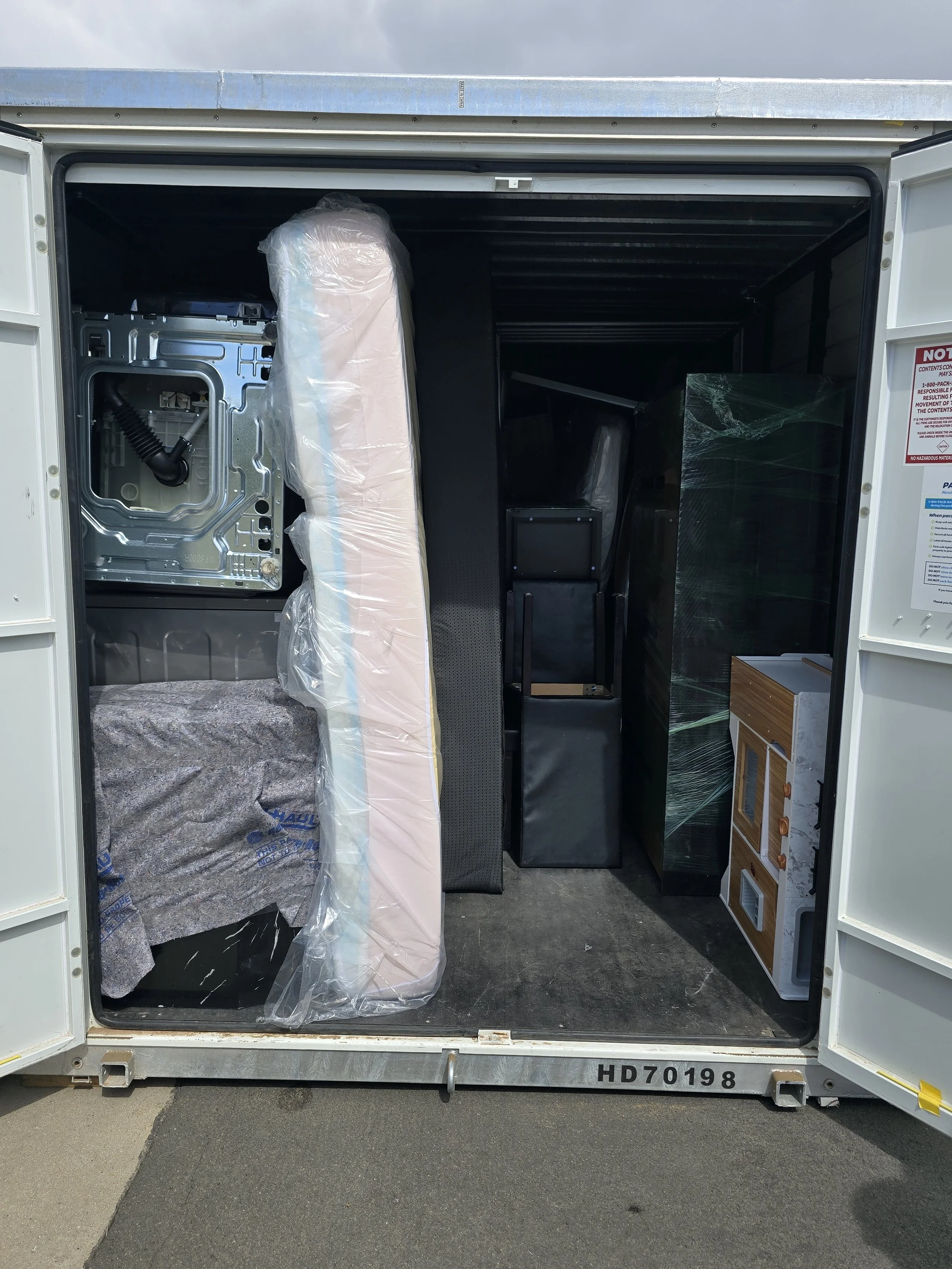 Open shipping container with various items including a wrapped mattress, black speaker, glass panel, small bookshelf, and plastic-wrapped items inside.