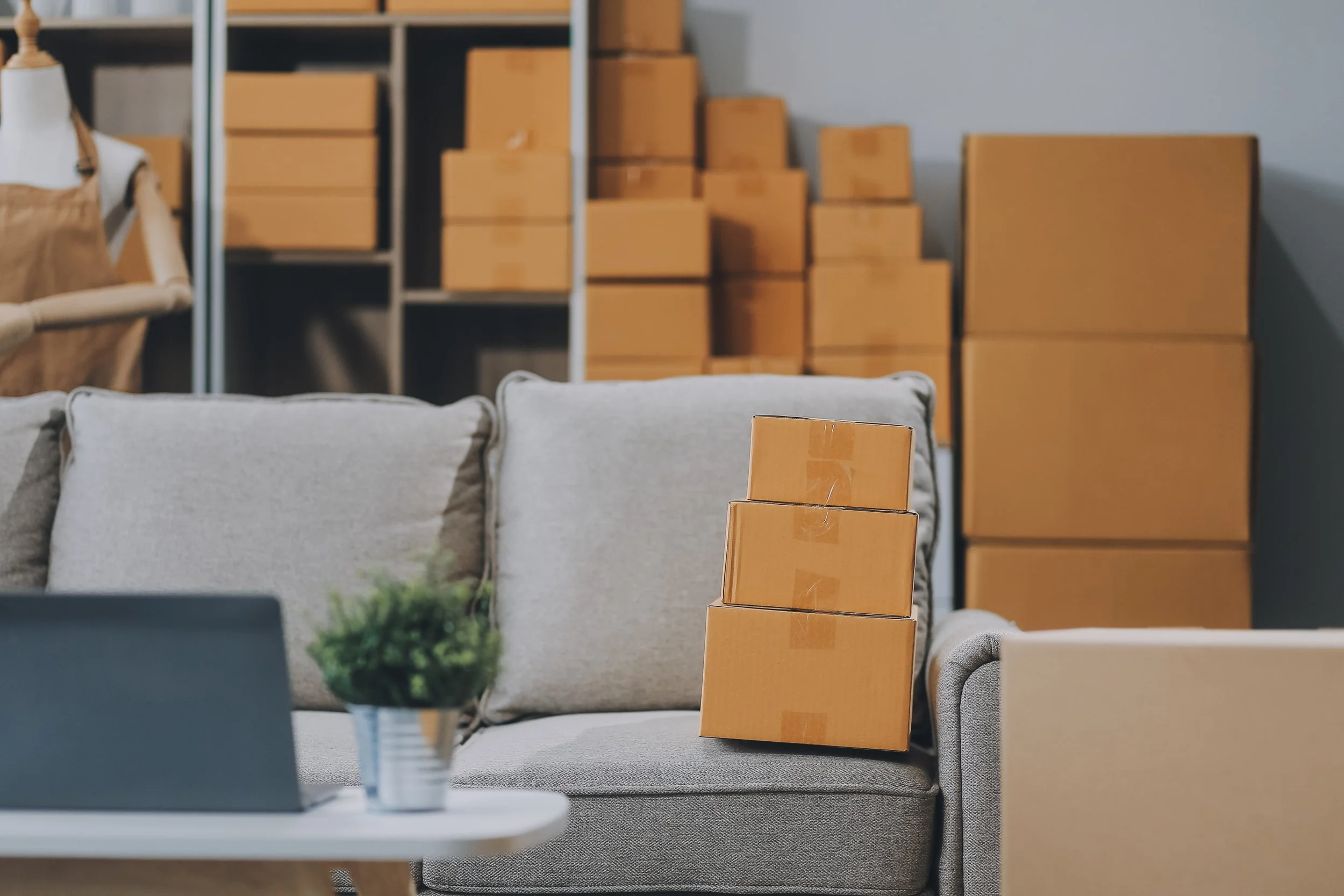 Stacked cardboard boxes on a gray sofa in a storage room with shelves filled with more boxes and a mannequin with clothing in the background.