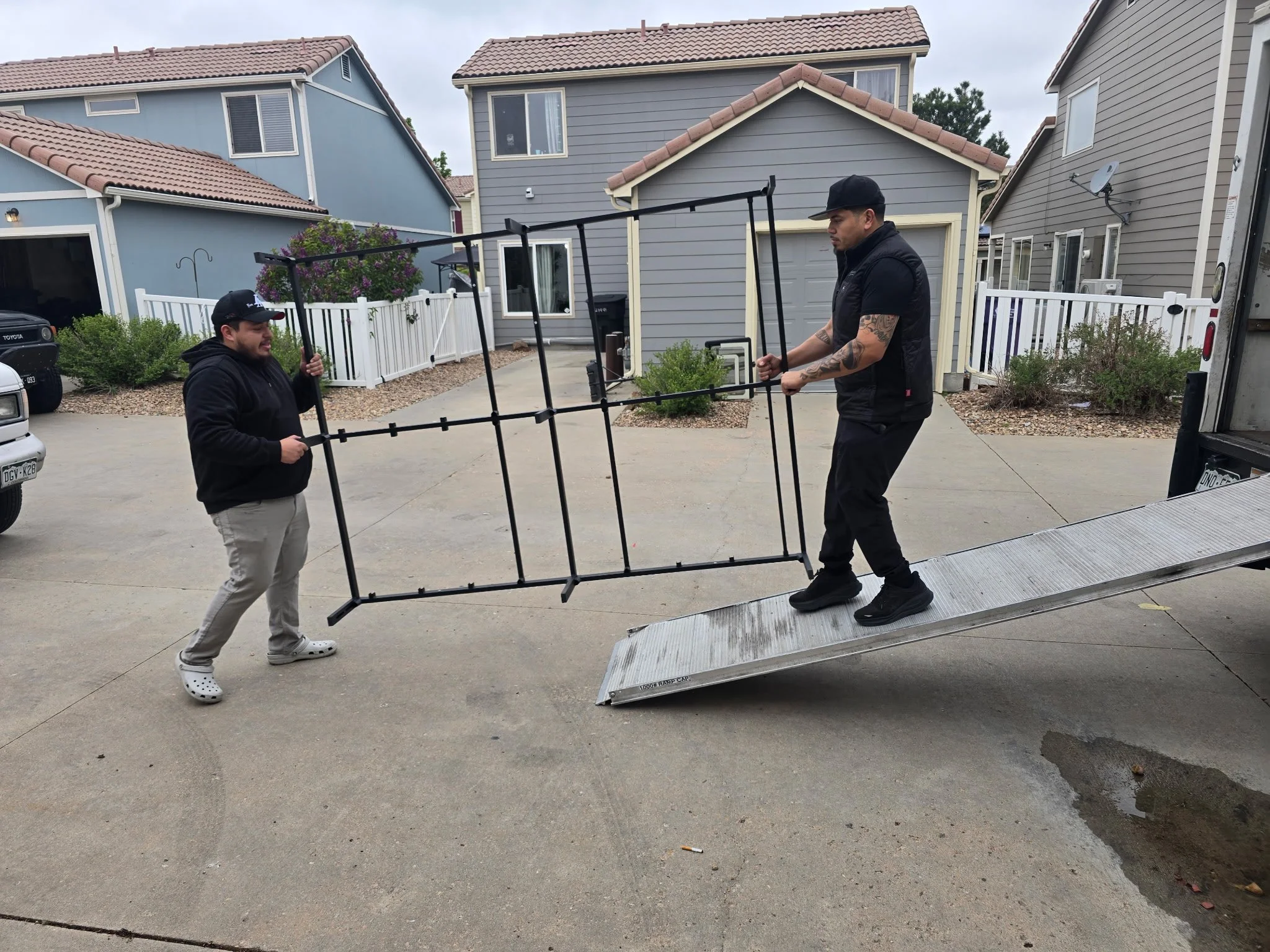 Two men are lifting a metal ramp onto a truck in a residential driveway, with houses and bushes in the background.