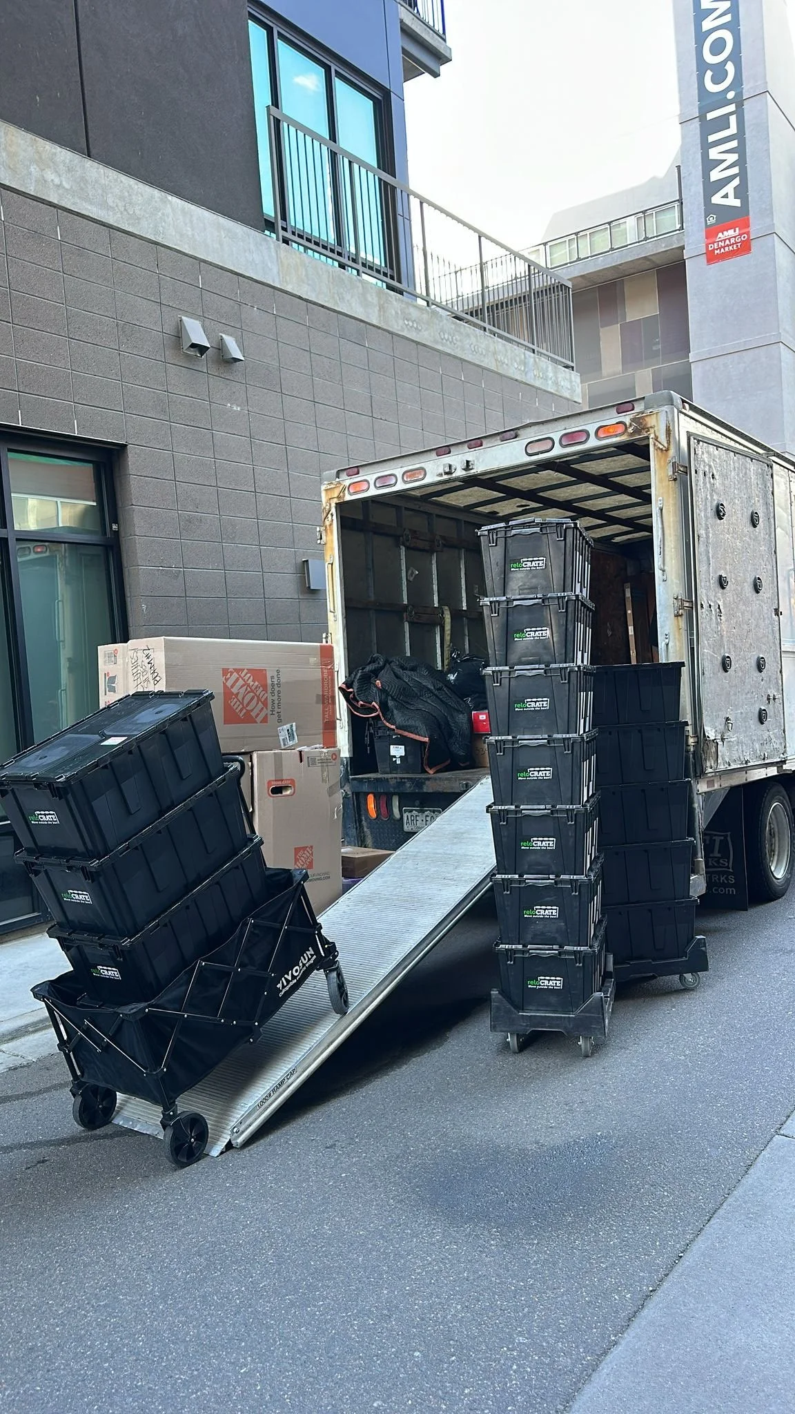 Moving truck with black plastic storage bins, boxes, and a ramp outside a modern apartment building.