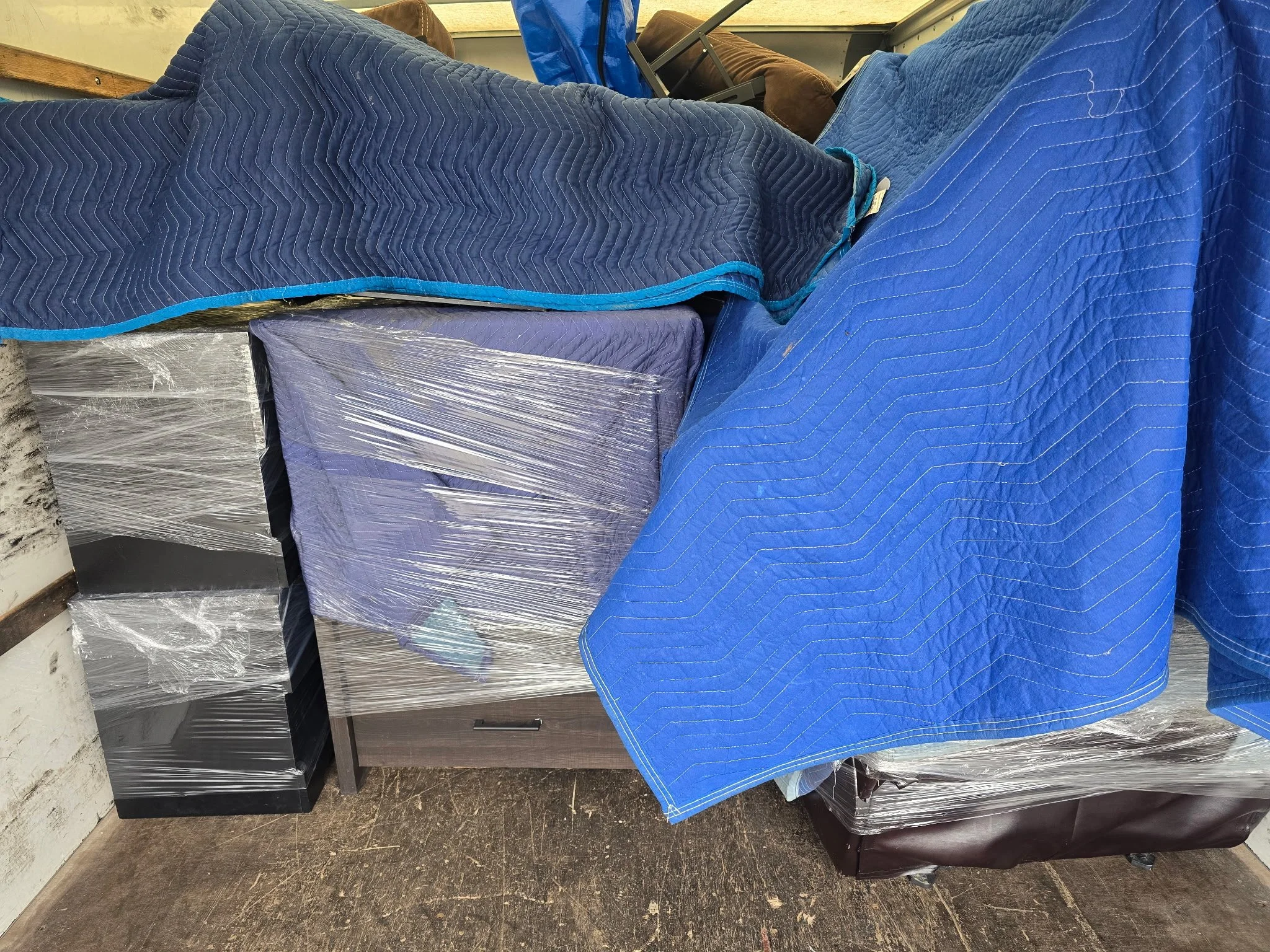 A storage room with couches covered in blue quilted fabric and plastic wrap, stacked against a wall, with wooden and metal furniture pieces visible in the background.