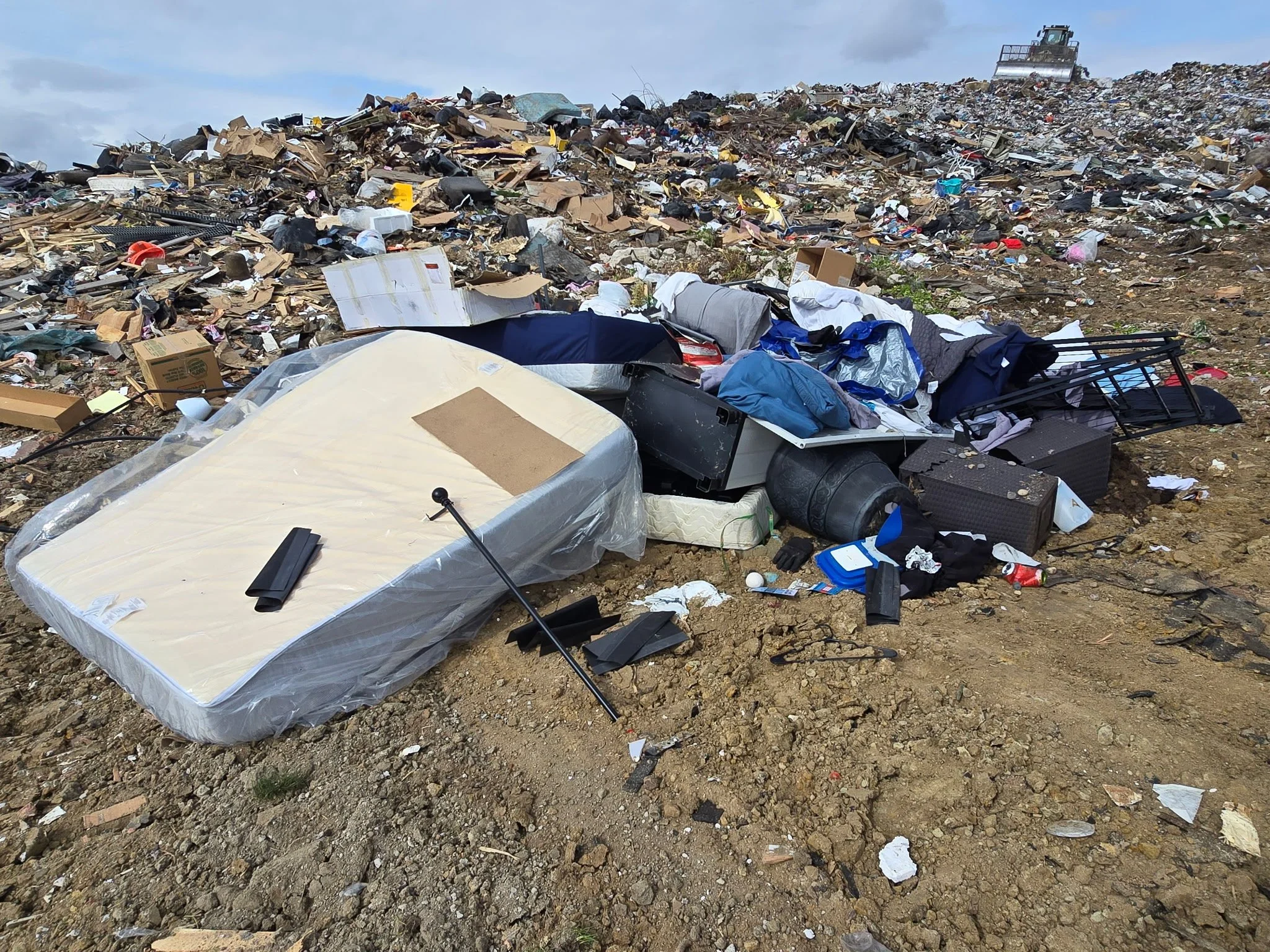 A large landfill filled with piles of trash, including cardboard, plastic, and discarded household items, stretching across a wide area with a bulldozer in the background.
