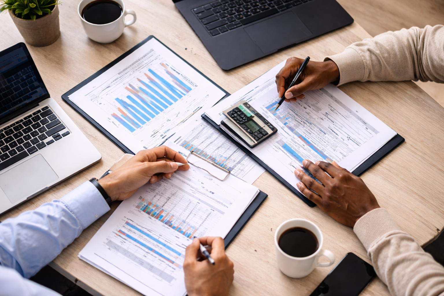 Two people analyzing financial graphs and reports at a wooden table, with laptops, a calculator, and coffee cups.