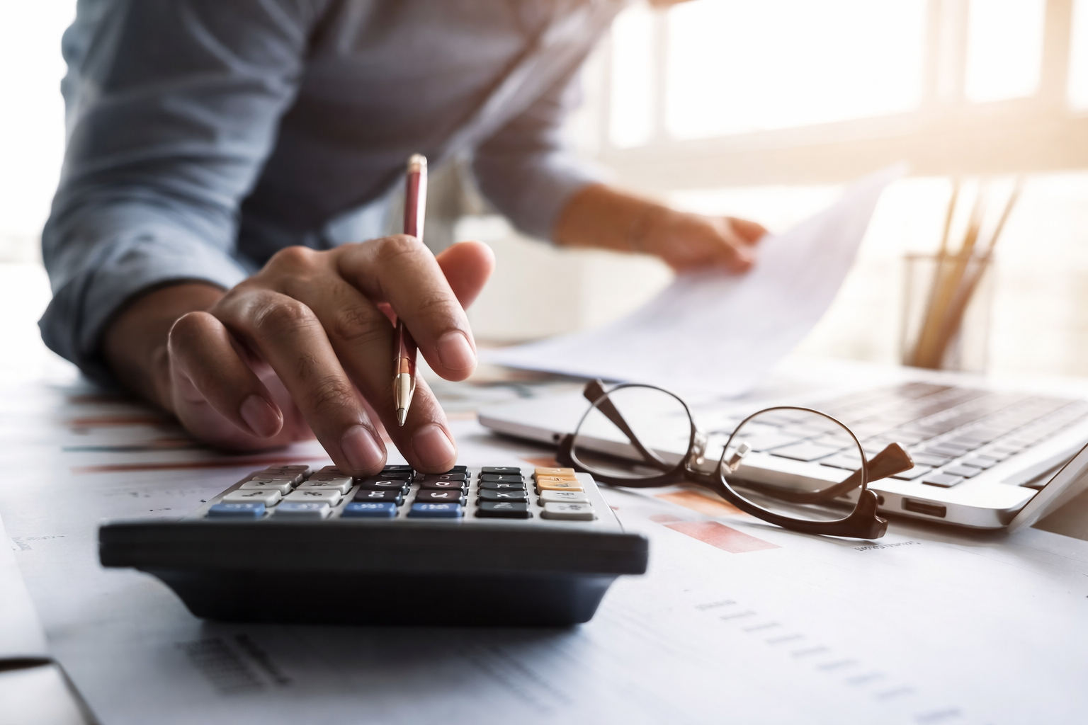 Person using calculator with documents, laptop, and glasses on desk in bright office.