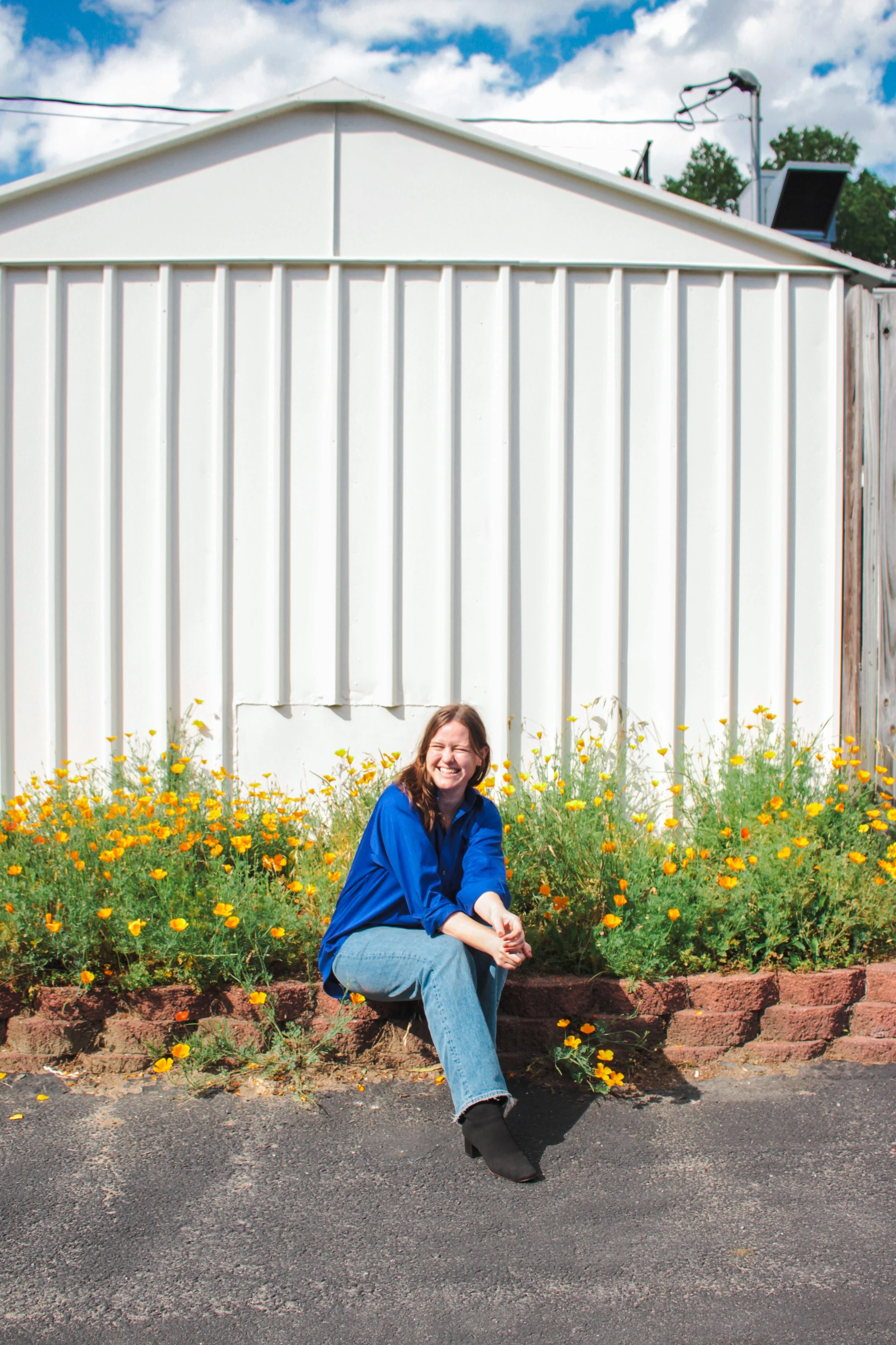 A woman with long hair, wearing a blue shirt, light blue jeans, and black shoes, sitting on a brick border beside yellow flowers in front of a white metal building, smiling at the camera.